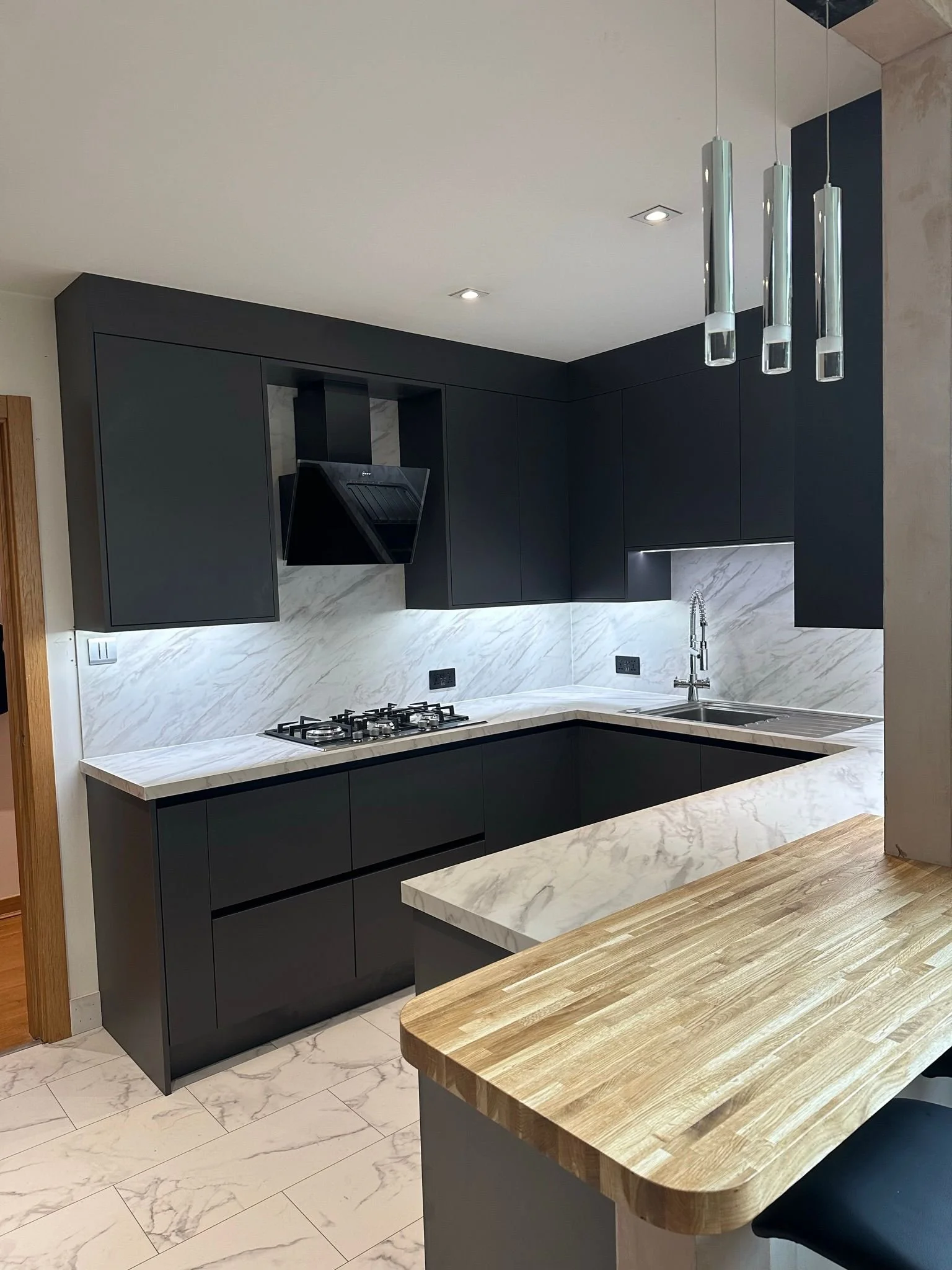 Modern kitchen with black cabinets, white marble countertops, and a wooden breakfast bar. Includes a stove, sink, and hanging pendant lights.