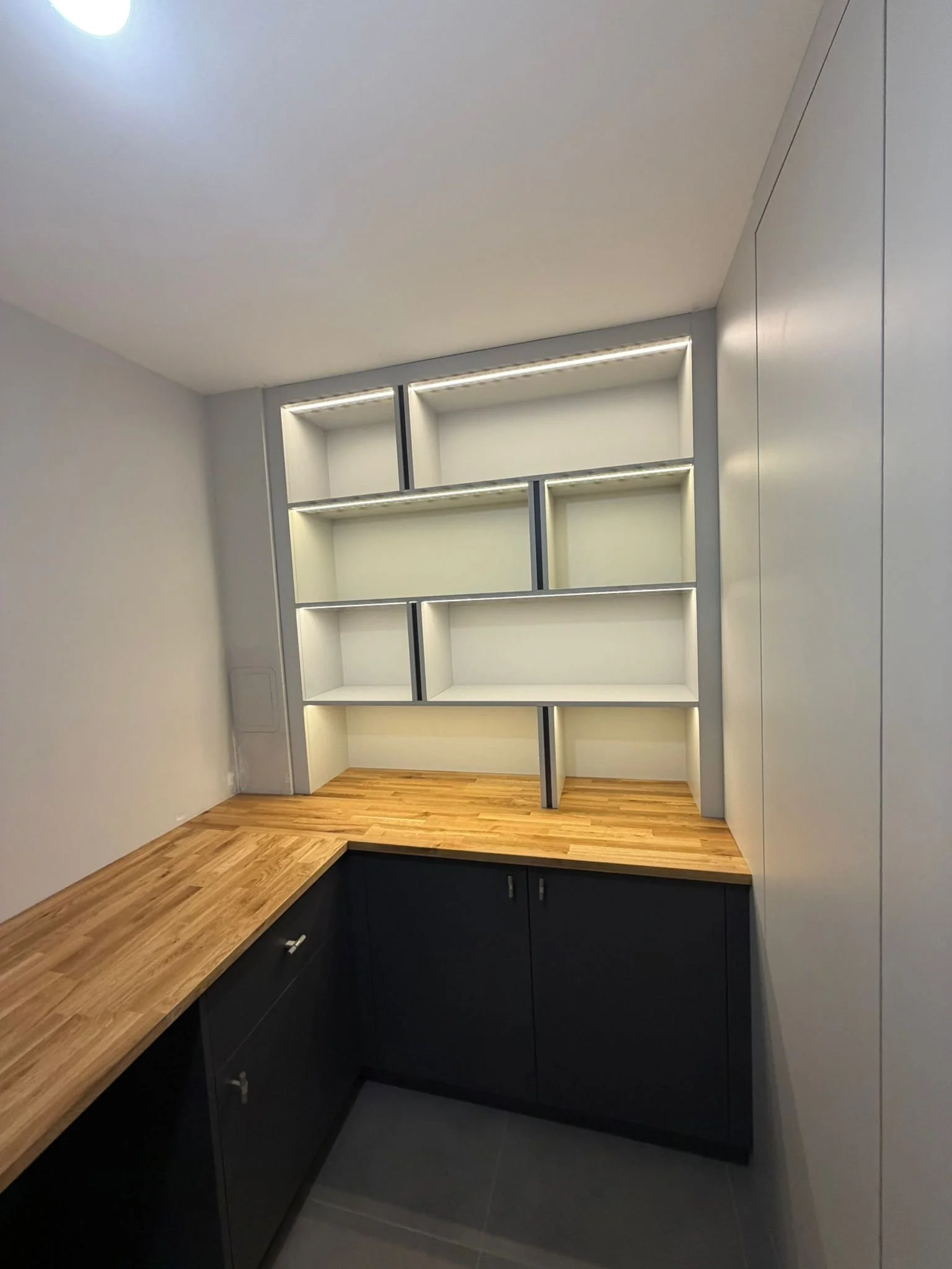 Modern built-in office with wooden countertop, dark grey cabinets, and white open shelves with integrated LED lighting.