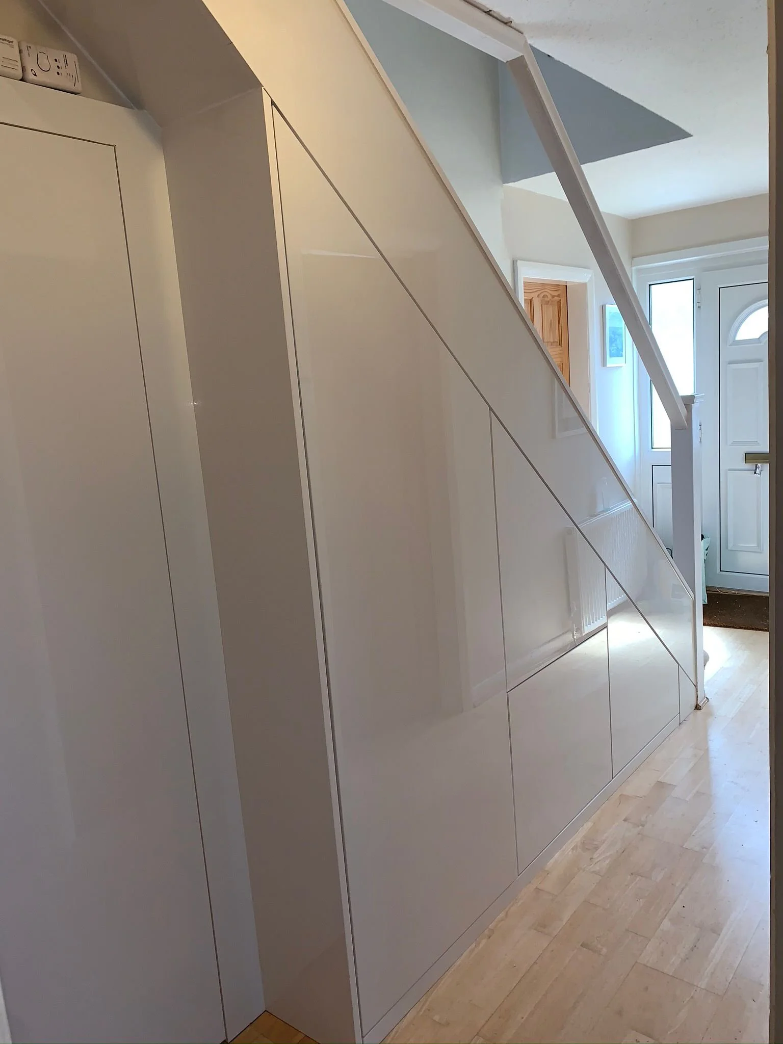 Modern, white, built-in storage unit under a staircase in a house entryway, with natural light coming through windows and a front door in the background.