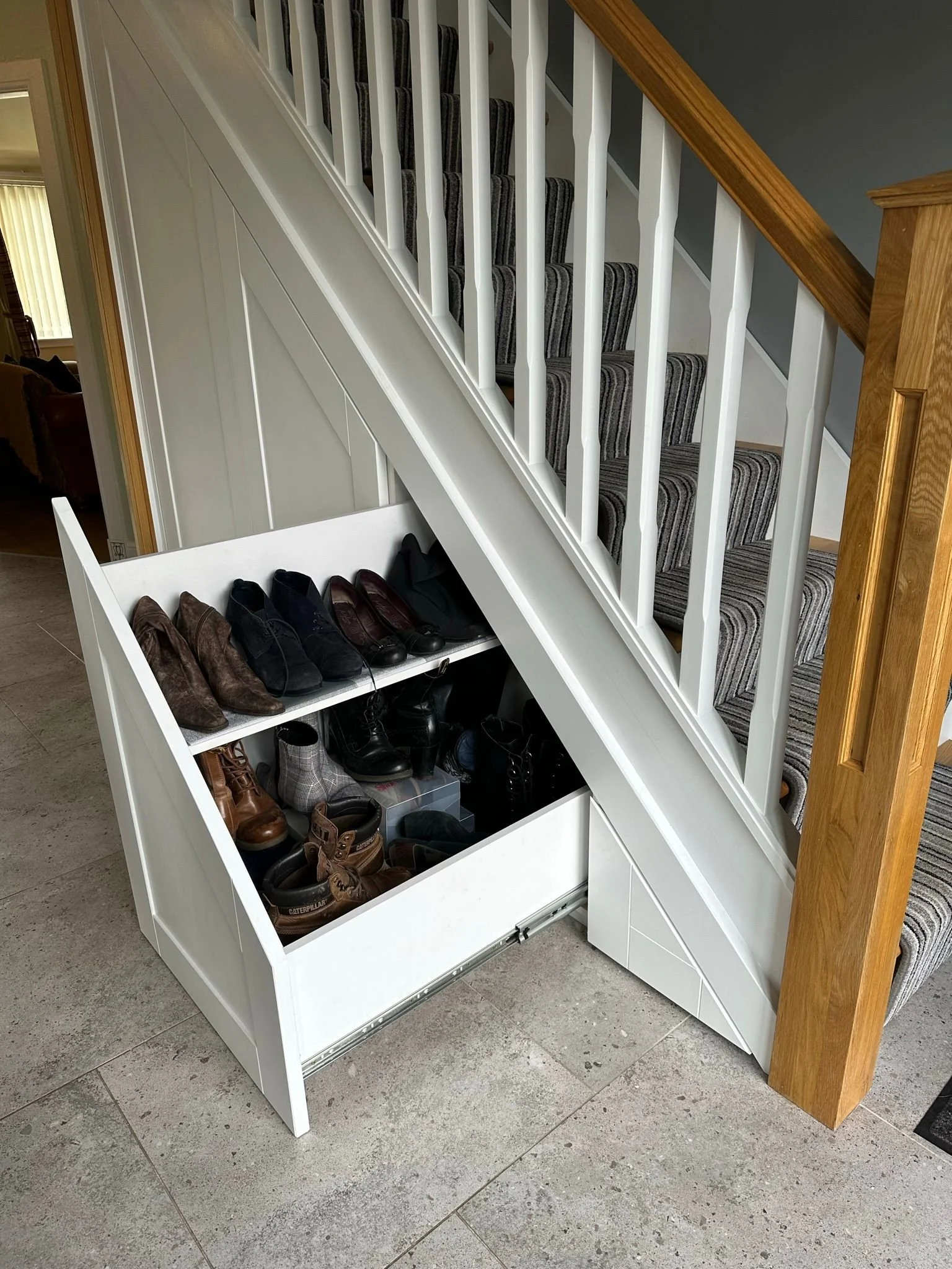 A white shoe storage bench filled with various shoes, placed at the base of a staircase with striped carpeted stairs and a wooden railing.