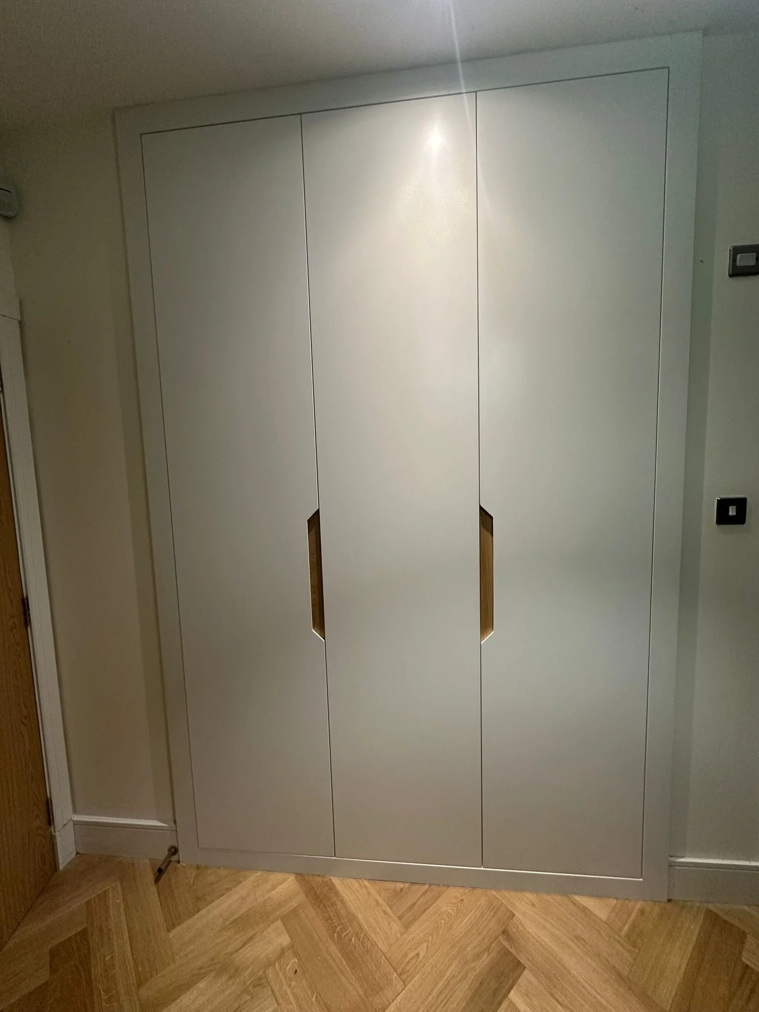 A white cabinet with three vertical panels, featuring two cut-out handles in the middle panels, located against a wall in a room with wooden floorboards.