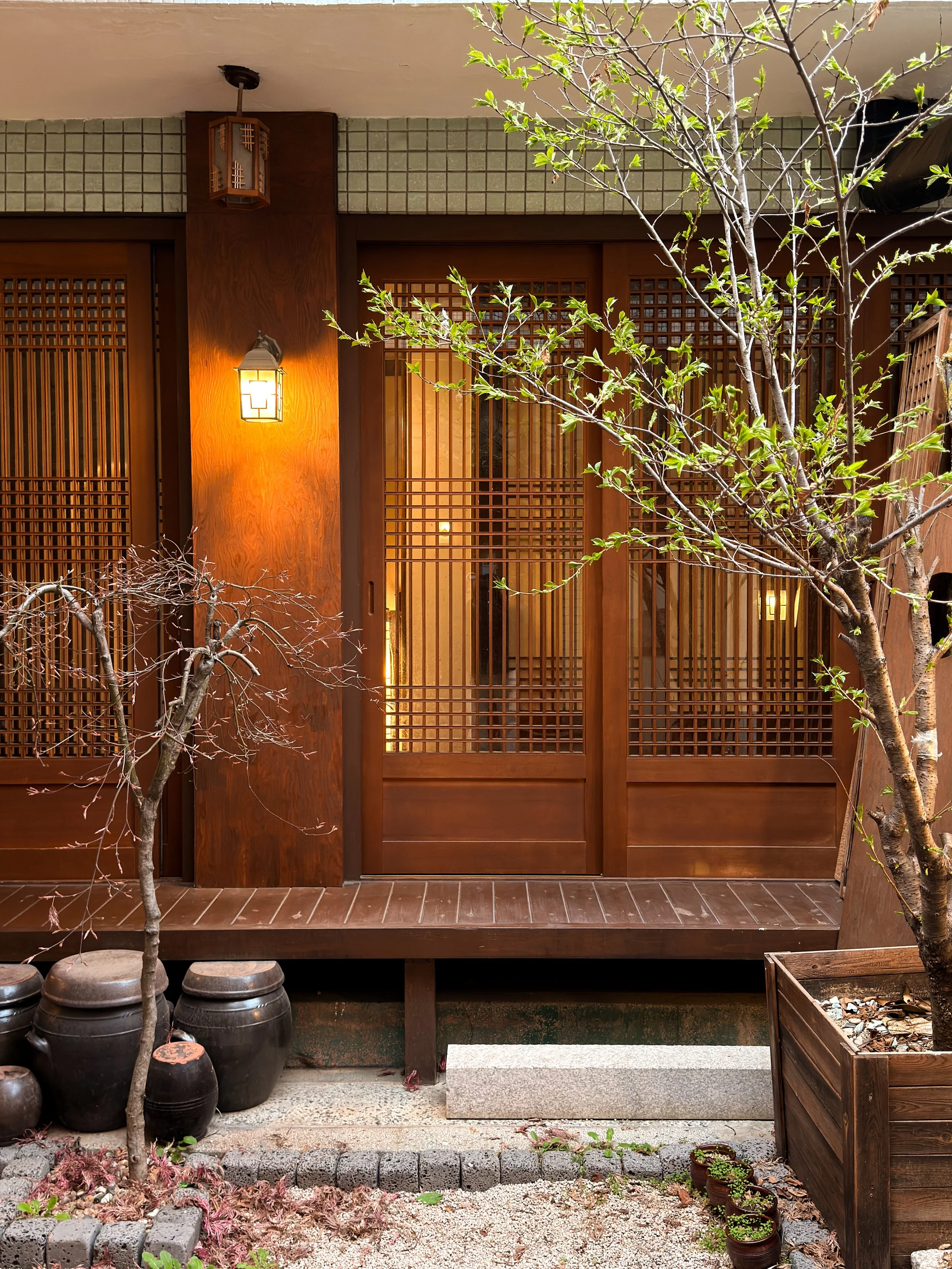 Traditional wooden door with lattice design, a warm wall-mounted lantern light, and two trees in planters in a courtyard.