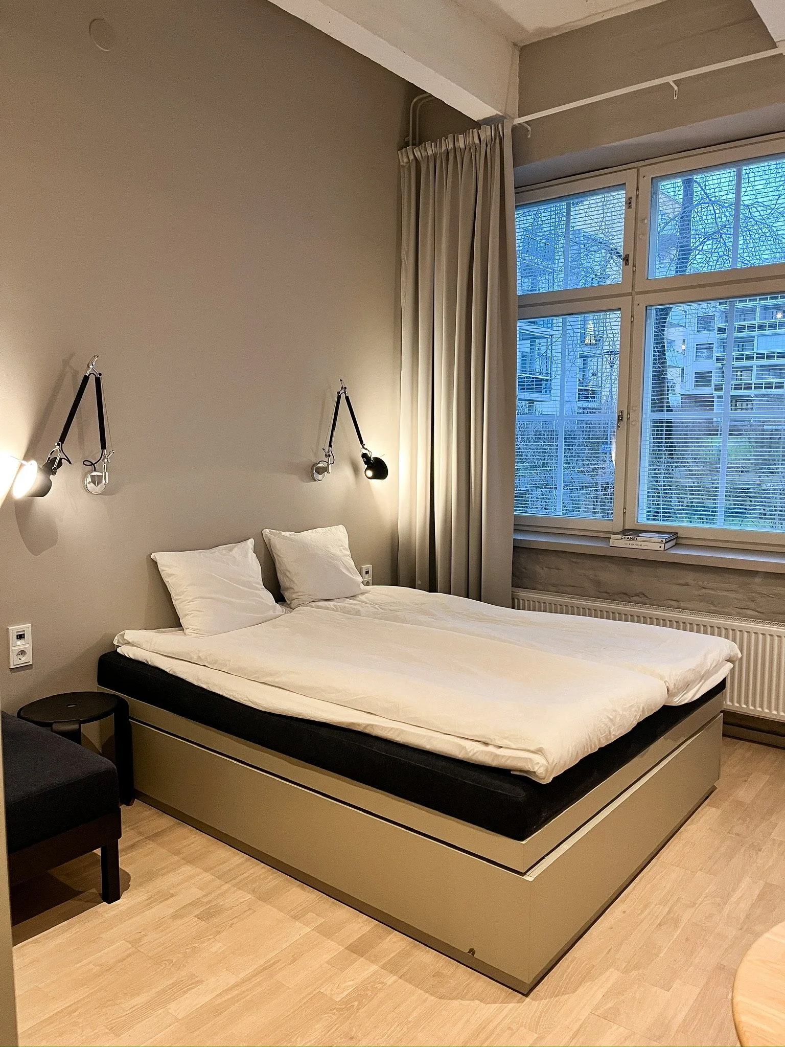 A neatly made bed with white pillows, black mattress, and beige bed frame in front of a grey wall. Two black adjustable reading lamps are mounted above the bed. To the right, there's a large window with white blinds and beige curtains, and through th