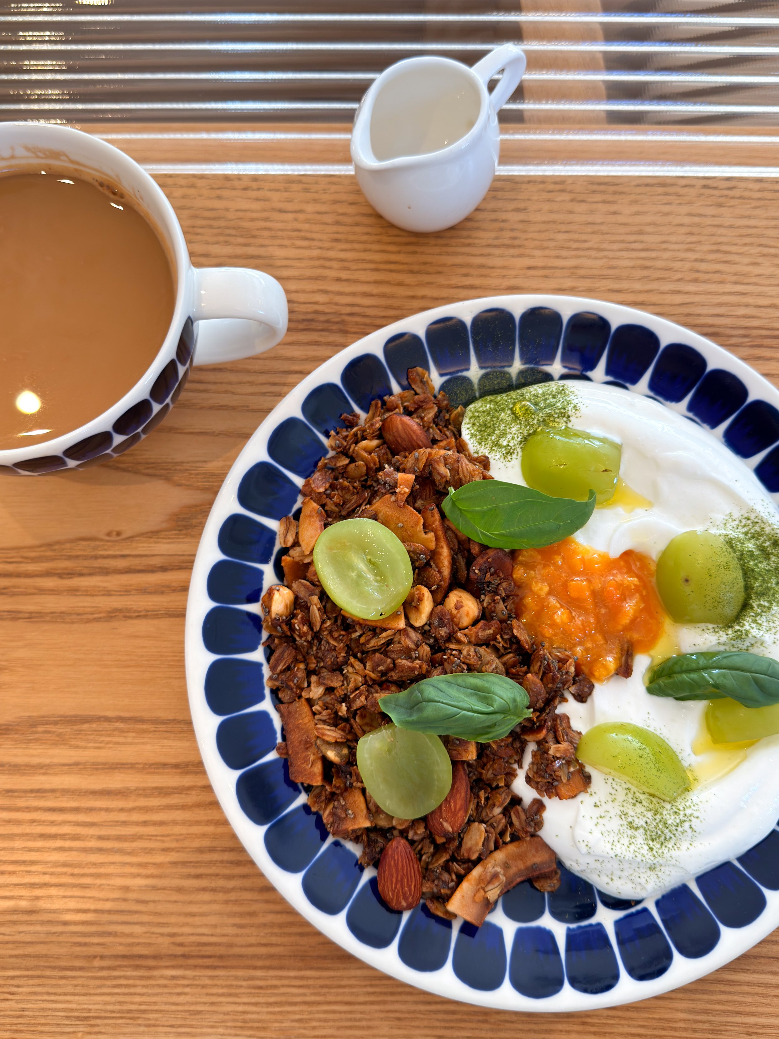 A breakfast plate with granola, green grapes, a fried egg, and herbs, served on a white dish with a blue pattern. A cup of coffee and a small pitcher of cream are also on a wooden table.