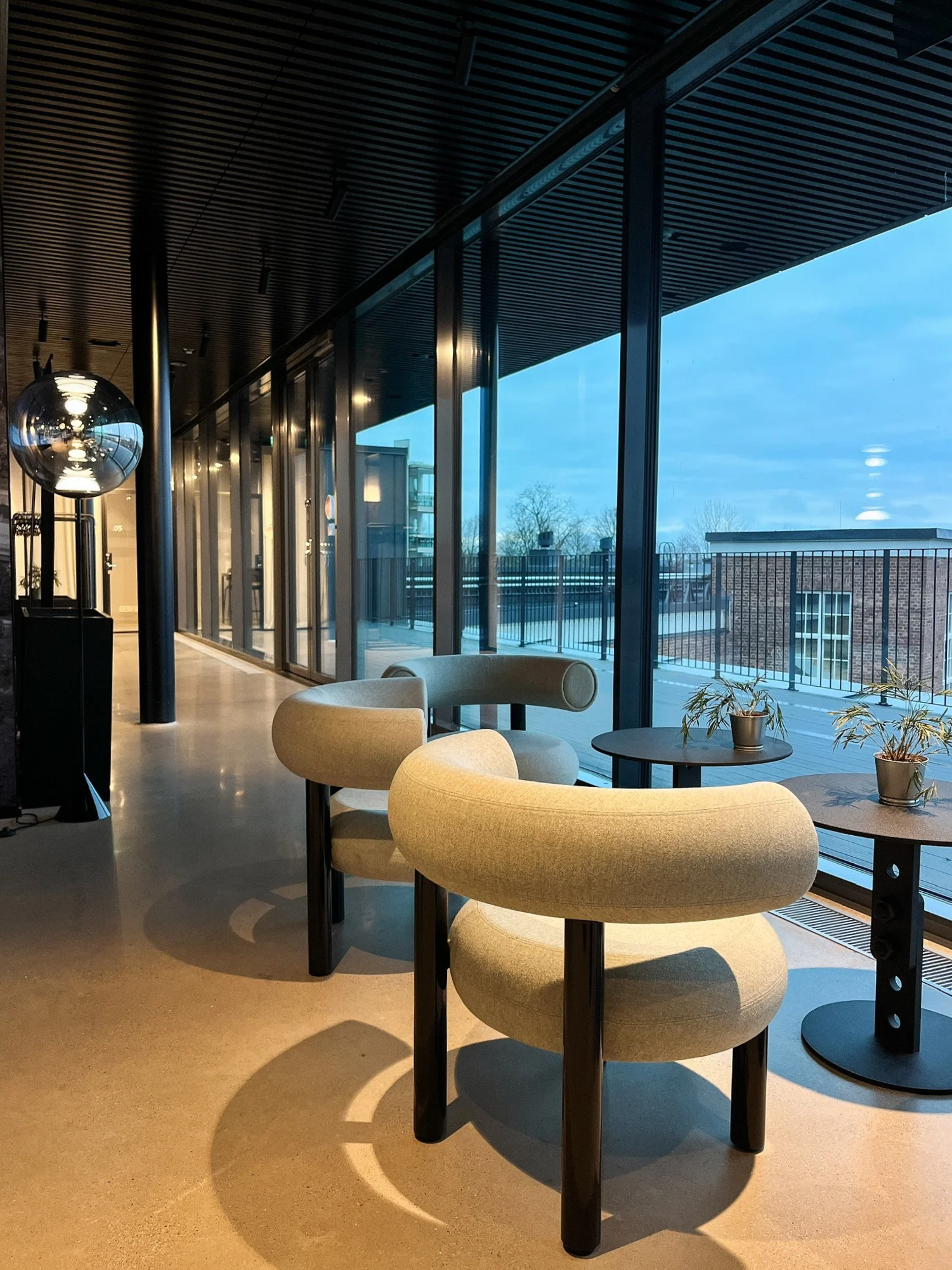 Modern interior with beige lounge chairs, small tables with potted plants, large floor-to-ceiling windows, and a wooden ceiling in a building with an urban view.
