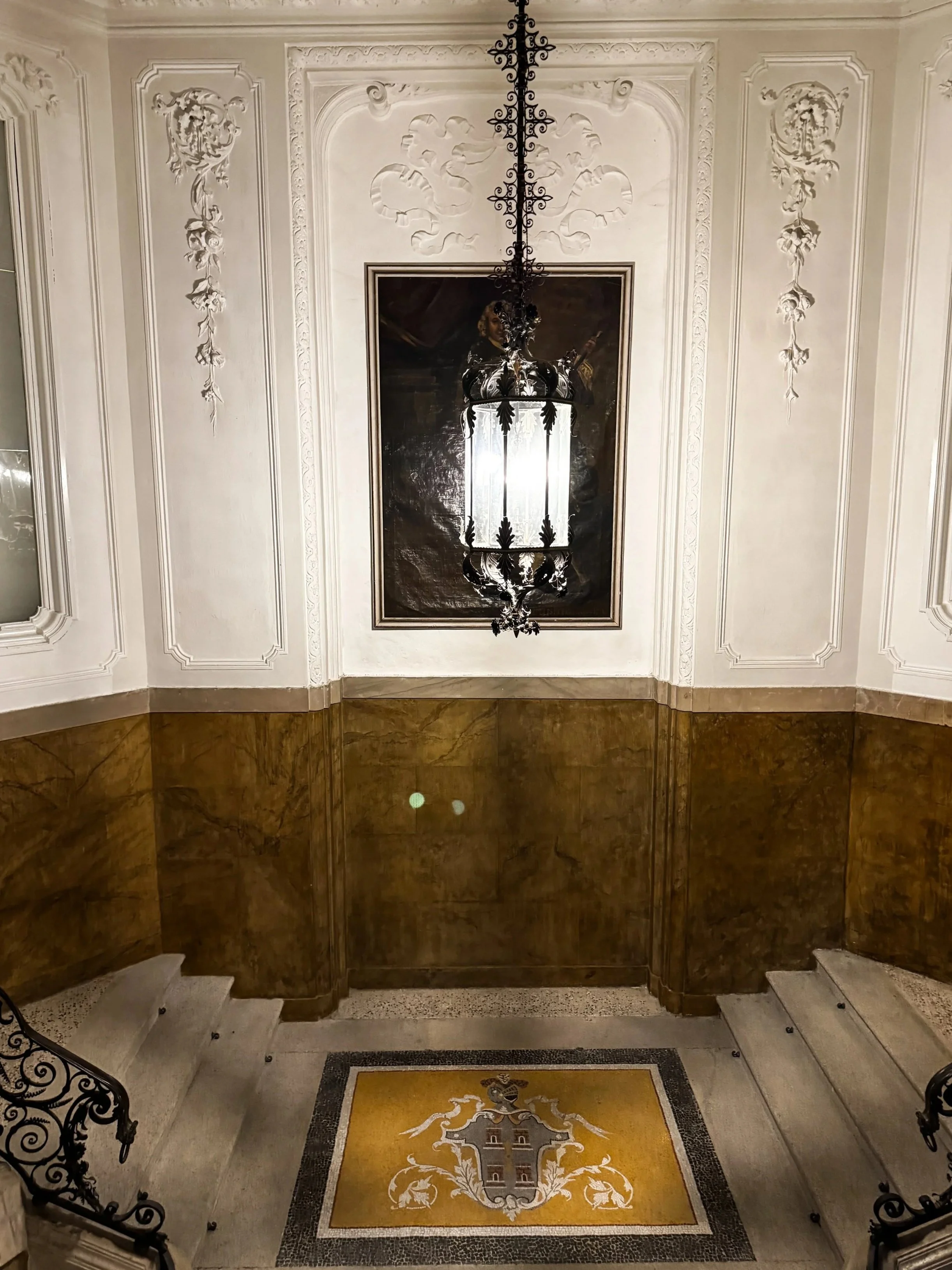 Elegant staircase entrance with ornate white walls, brown marble paneling, a decorative heraldic floor tile, a large framed portrait, and an intricate hanging lantern.