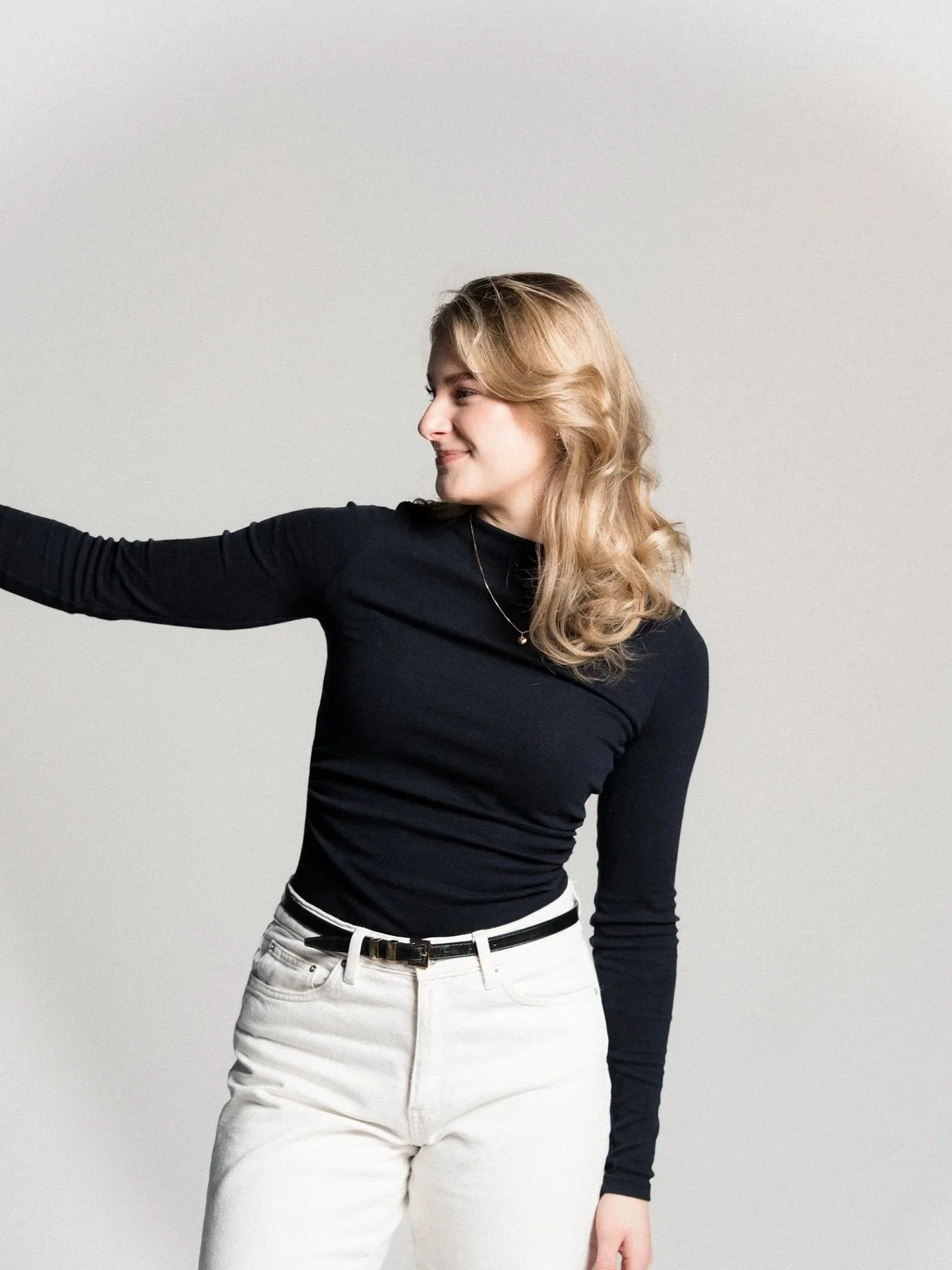 A woman with wavy blonde hair wearing a black long-sleeve shirt and white pants, standing against a neutral background, looking to her left.