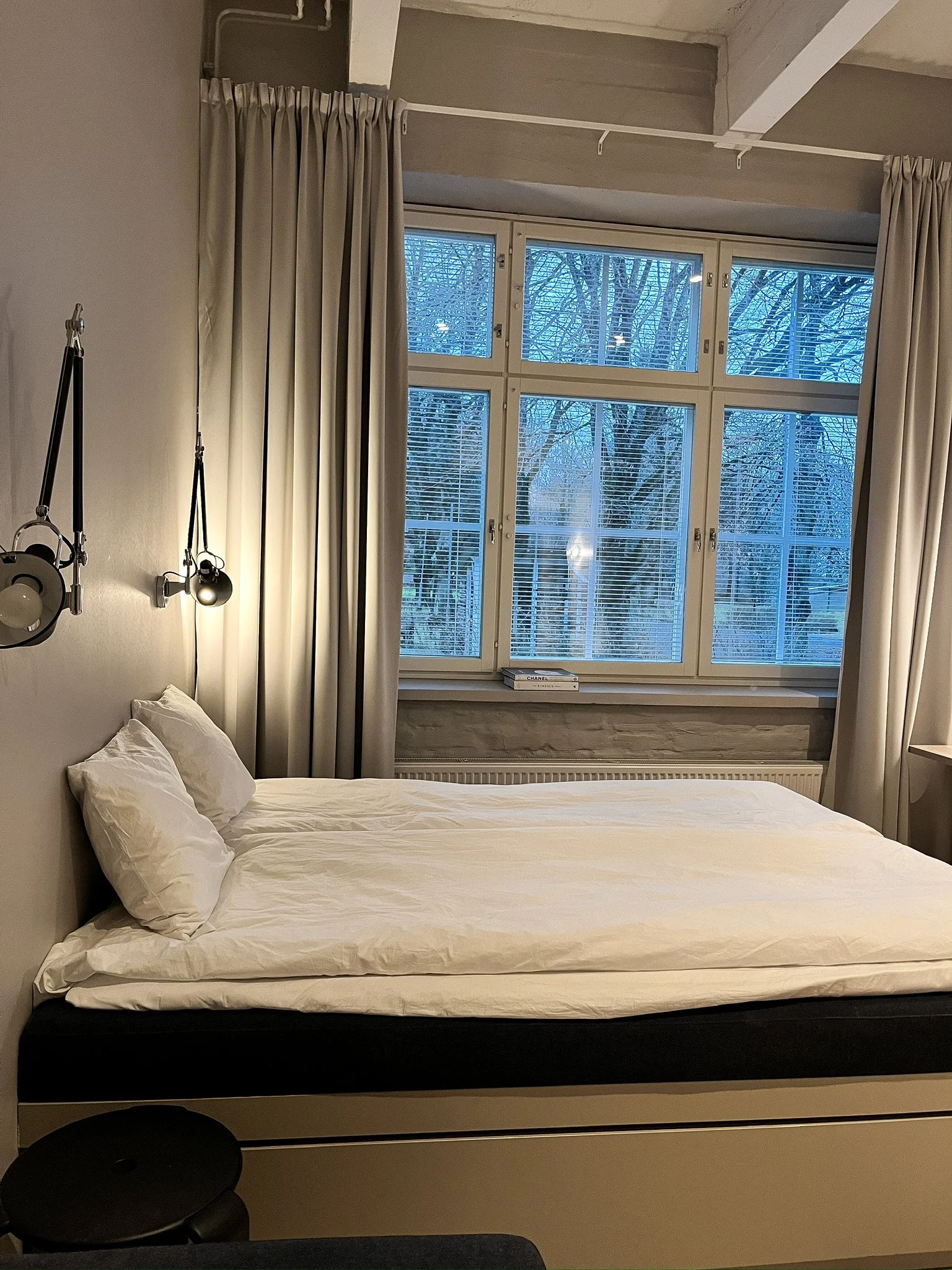 A neatly made bed with white sheets and pillows in a modern bedroom. There are wall-mounted lights on each side of the bed. The window behind the bed shows a view of leafless trees outside, with horizontal blinds and cream curtains.
