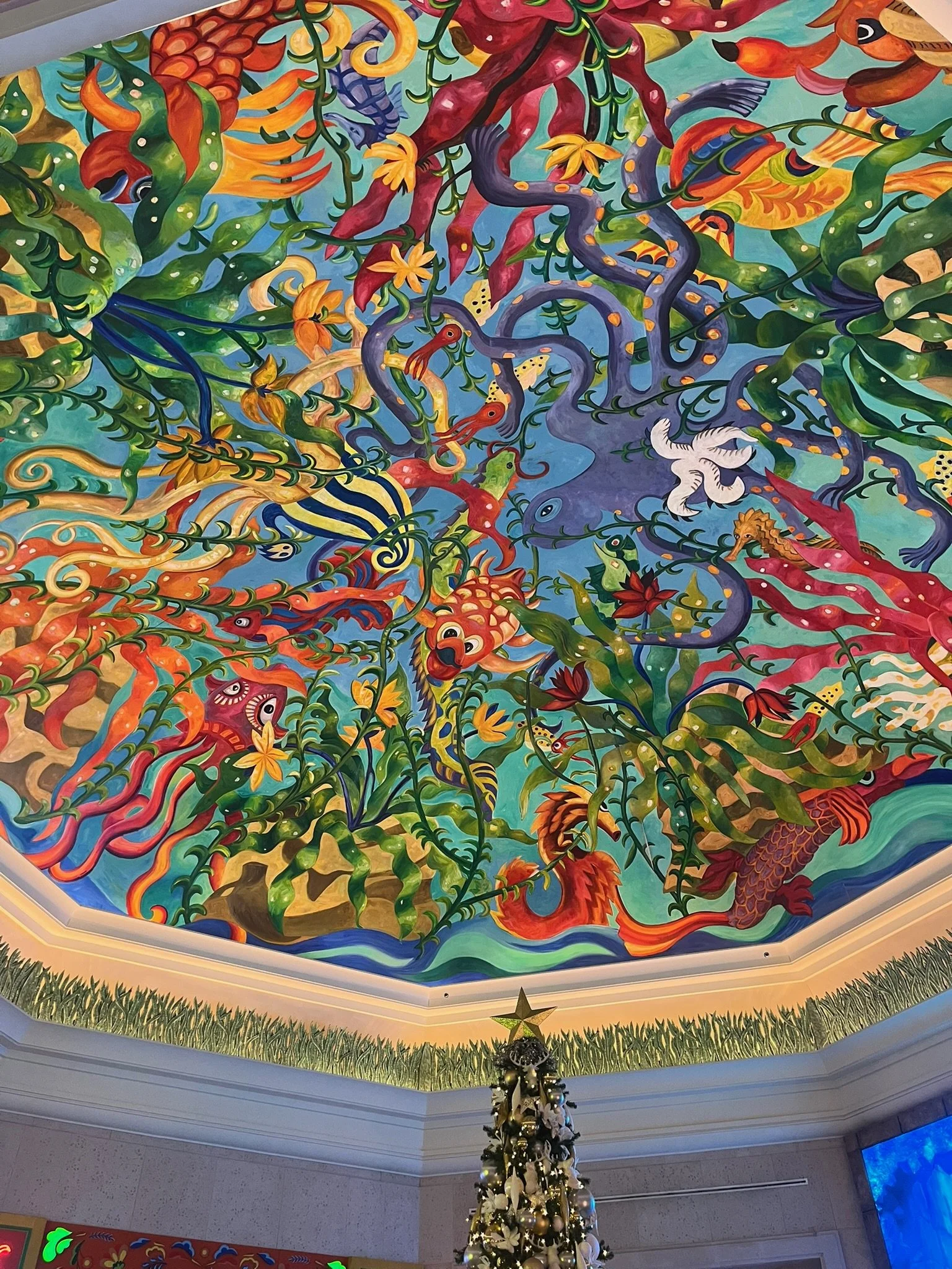 Colorful mural painting on ceiling featuring various stylized mythical creatures, plants, and flowers in vibrant colors, with a decorated Christmas tree below.