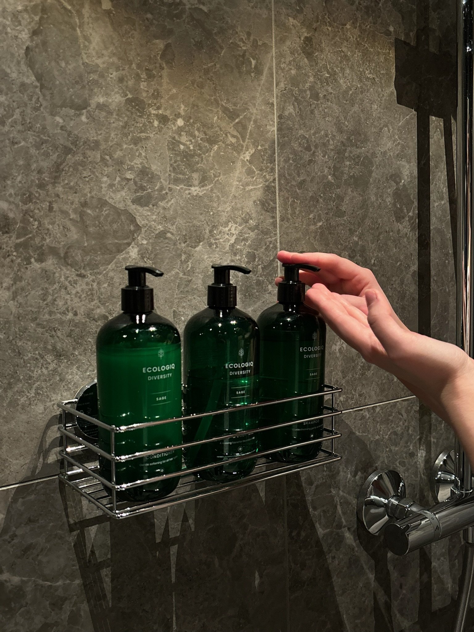 A person's hand reaching to grab a green pump bottle of shampoo in a bathroom shower with gray marble tiles, along with two other green bottles of conditioner and body wash in a wire shelf.