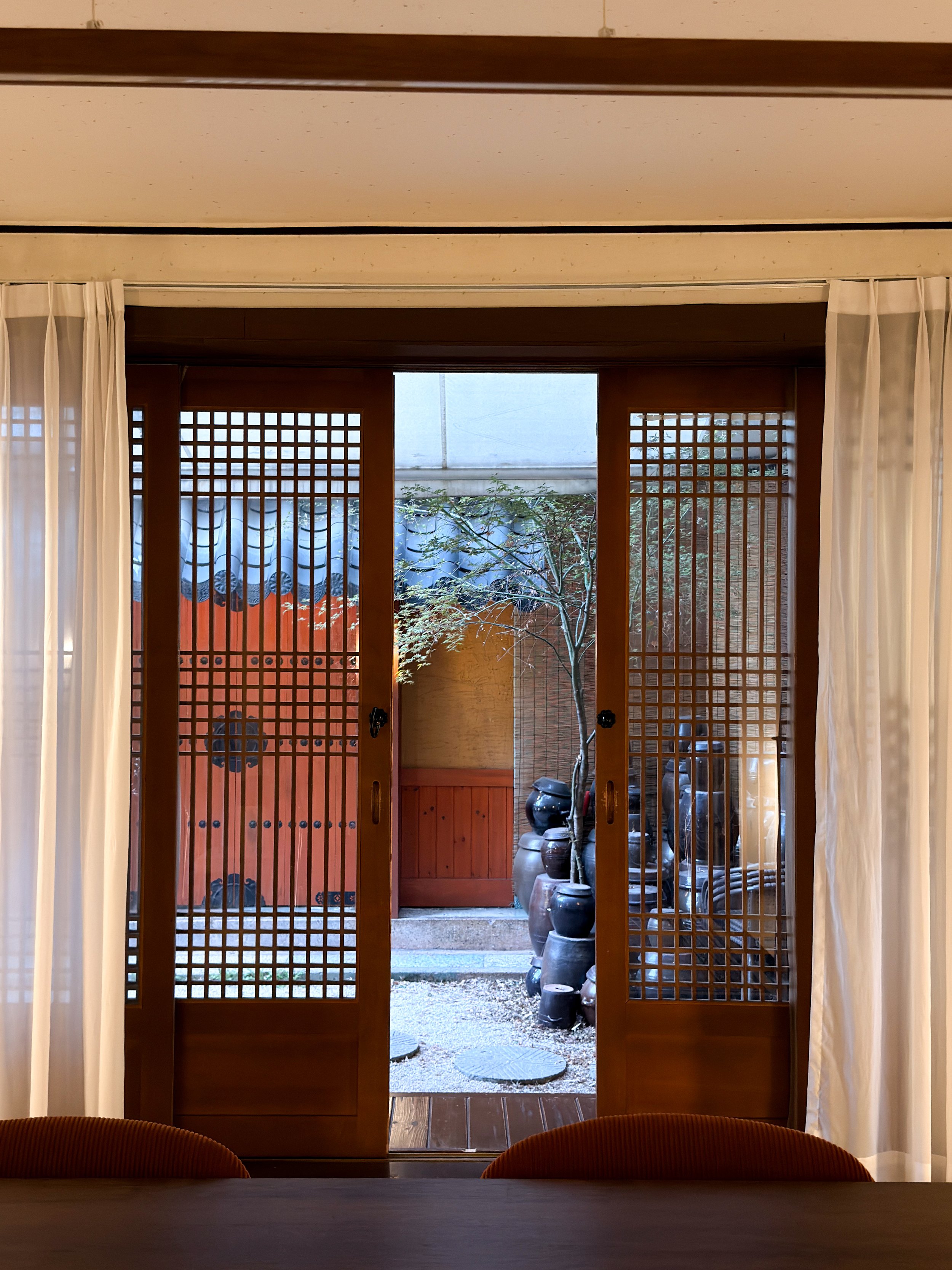 View through open wooden sliding doors to a small outdoor garden with clay pots, a small tree, and a red wooden wall.