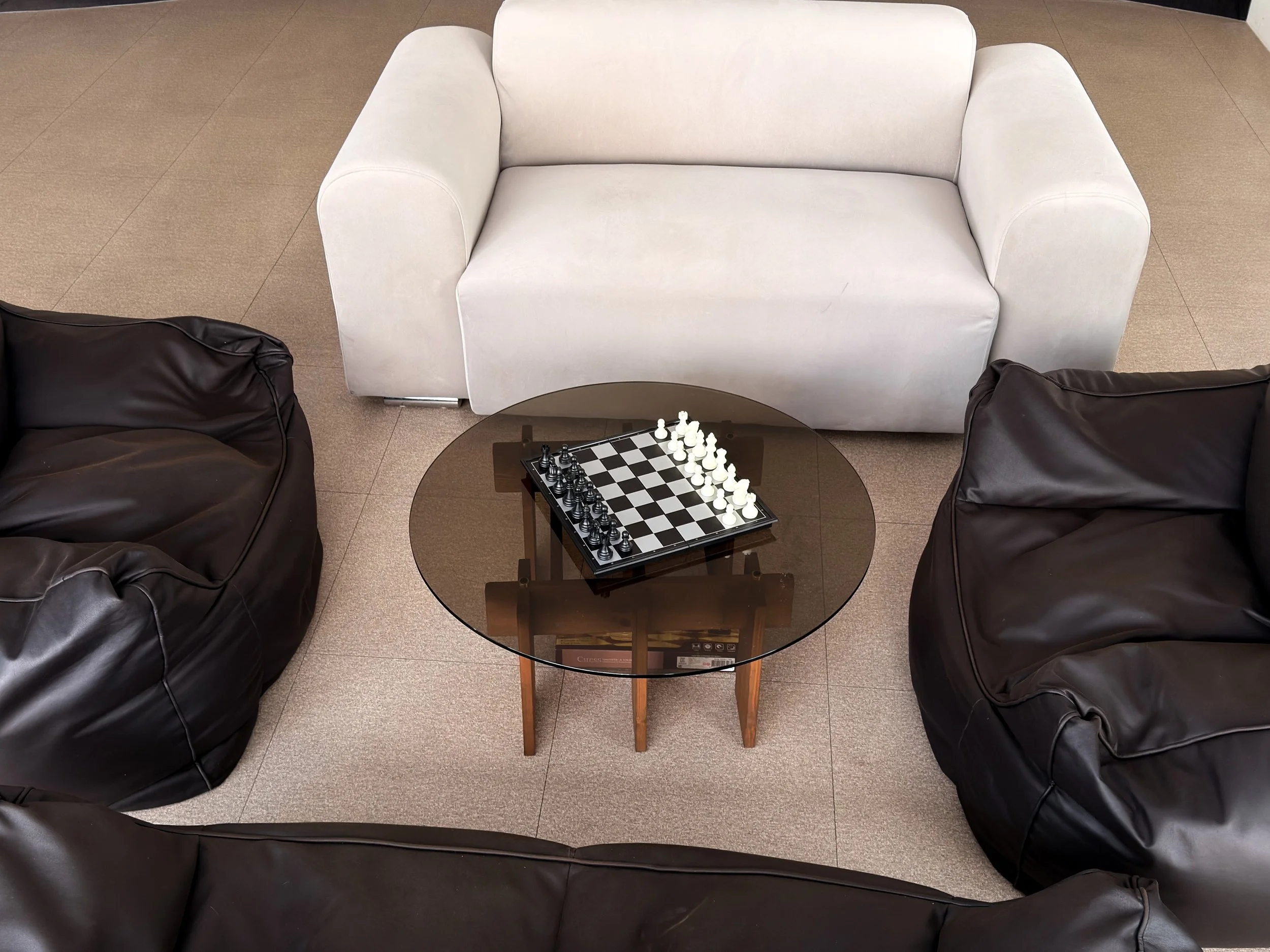 A seating area with three black leather armchairs arranged around a round glass coffee table. A chessboard is set up on the table, with white pieces in starting positions.
