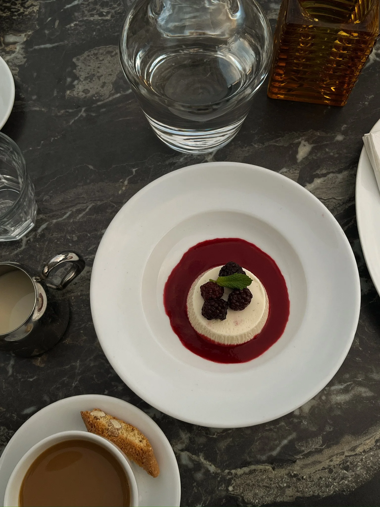 A dessert of panna cotta with blackberries, raspberry sauce, and a mint leaf on top, served on a white plate. Surrounding the dessert are a glass of water, a cup of tea with a biscotti, an empty small pitcher, a glass of clear water, and a brown sugar or syrup dispenser on a dark marble table.