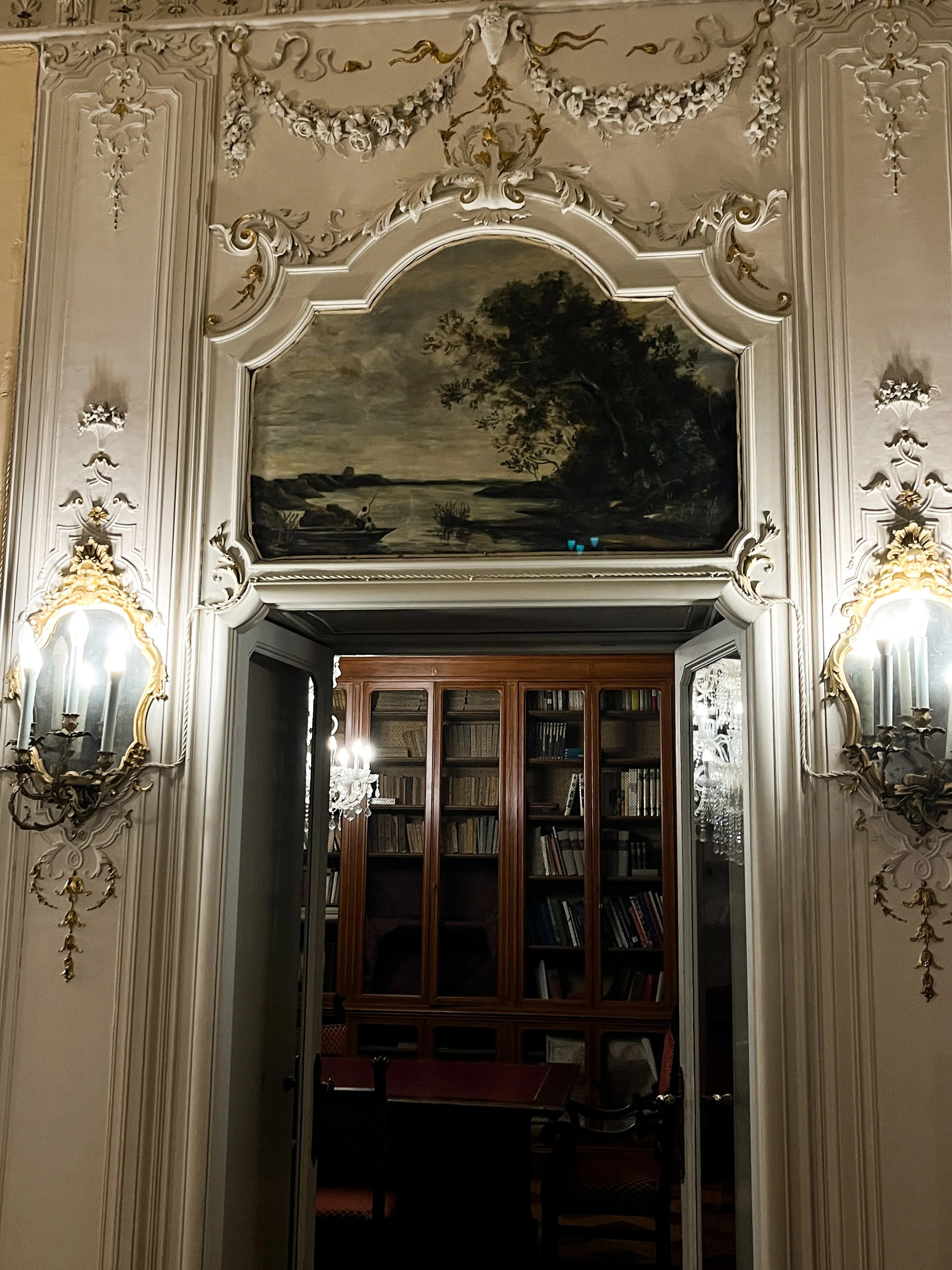 Elegant room with tall wooden bookshelves filled with books, ornate cream-colored walls with gold accents, framed landscape painting above the doorway, and mirrors with gold trim and wall sconces with candles.