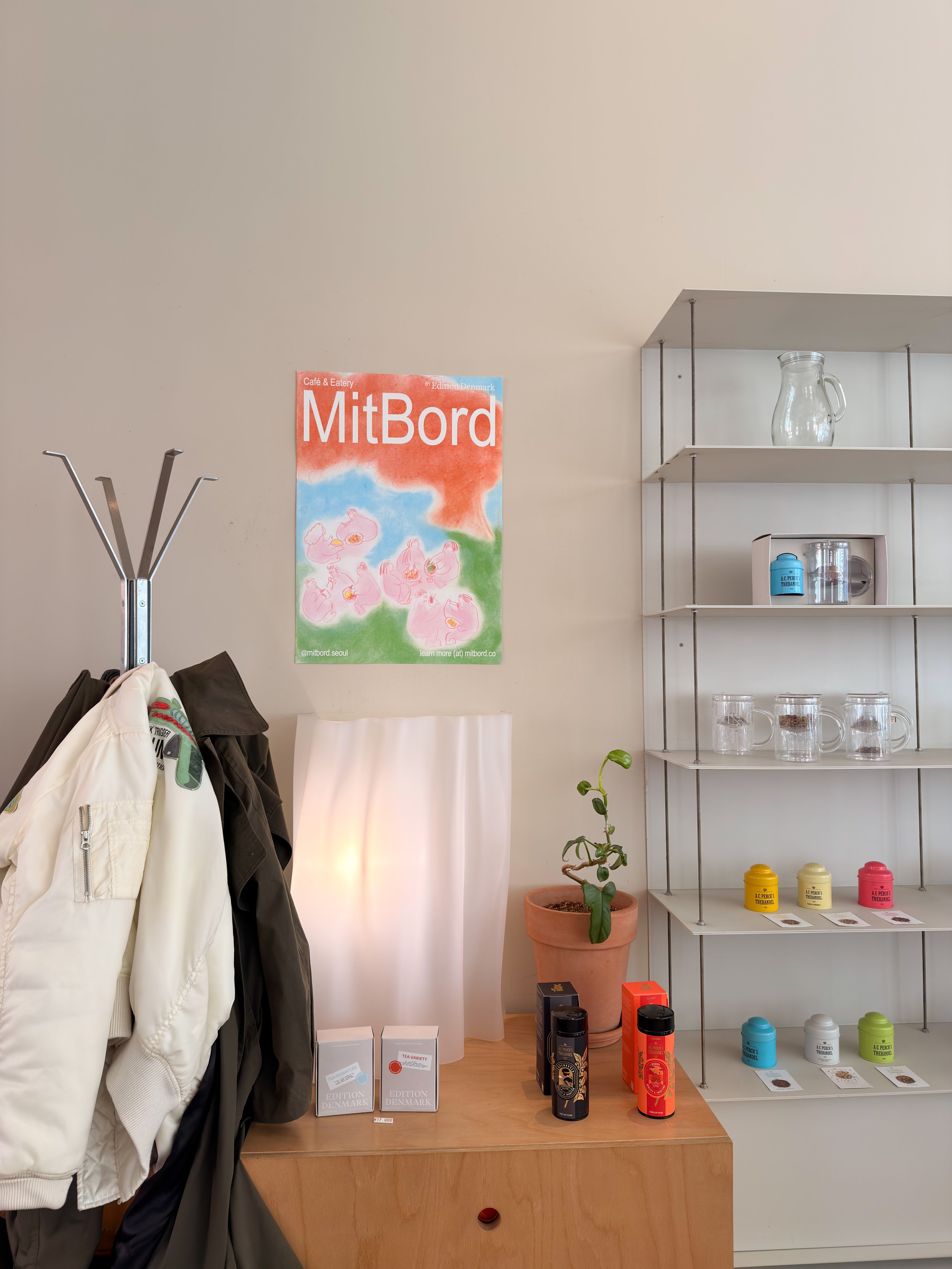 Interior of a room with hanging jackets, a poster with the word 'MitBord', a wooden cabinet with boxes and bottles, a potted plant, and a white shelf with glass jars and containers.