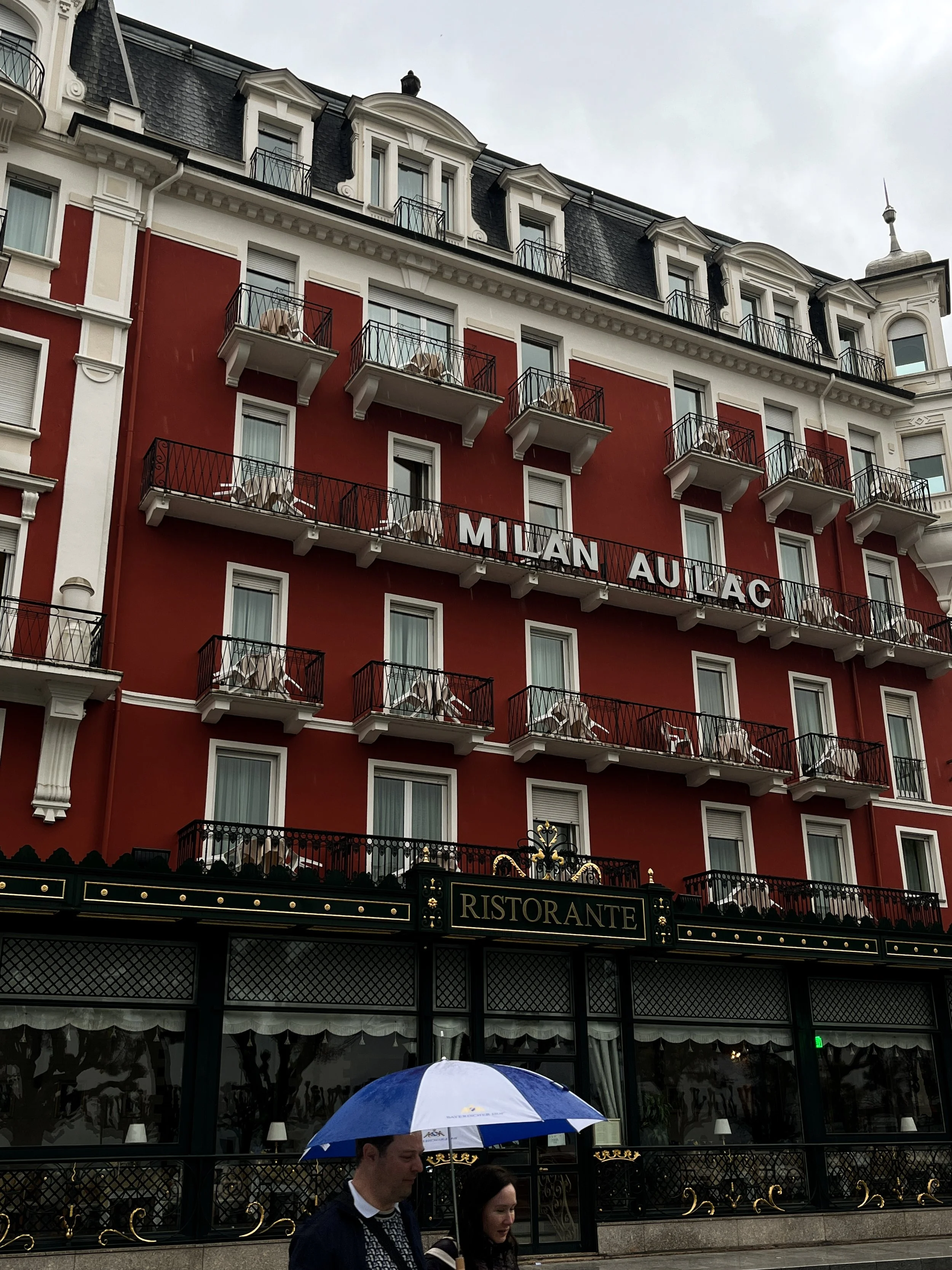 A European-style hotel building with balconies, a sign reading 'Milan Au Lac,' and a restaurant labeled 'Ristorante' at ground level. Two people walking with an umbrella in front of the building.