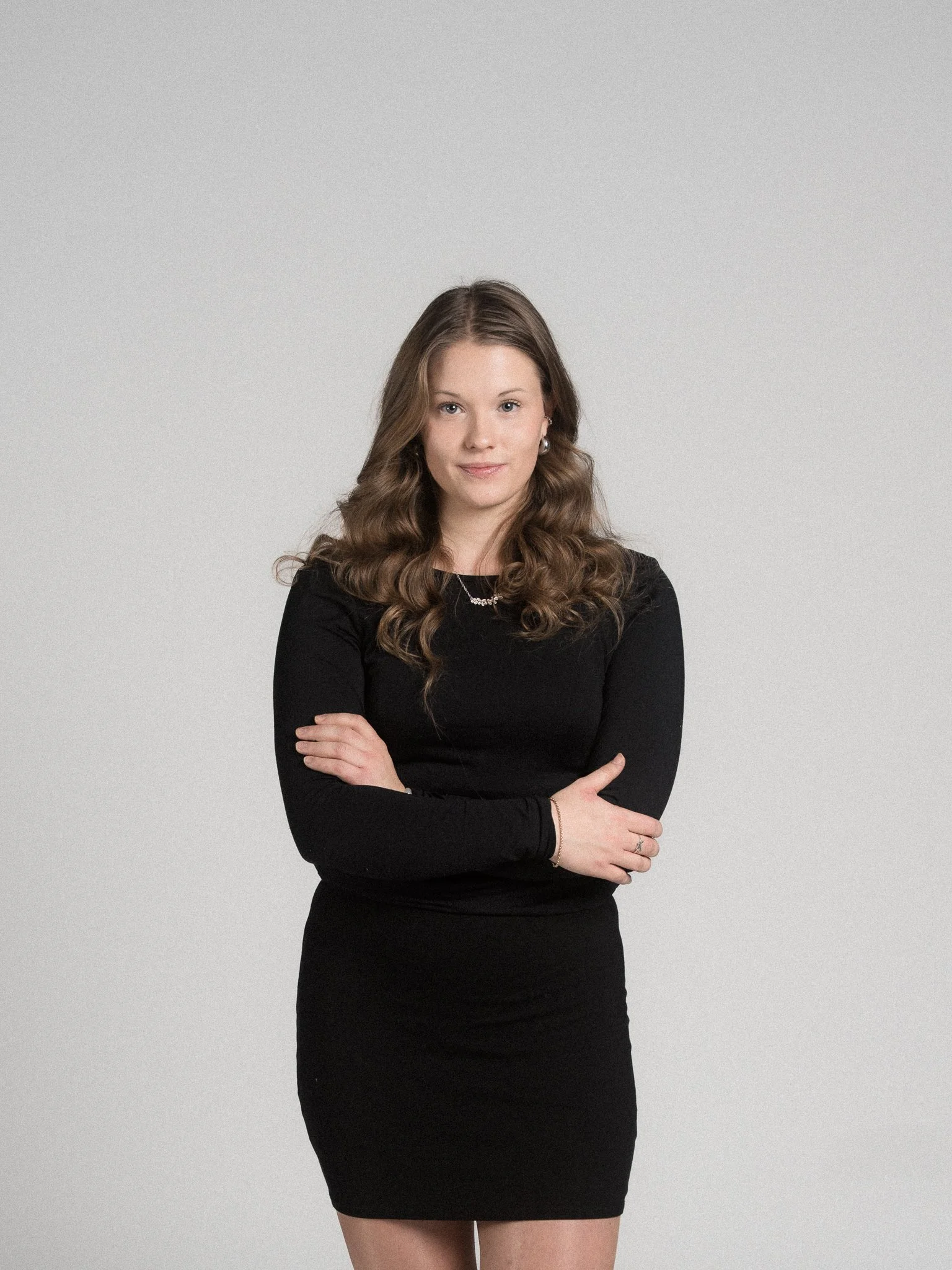 A woman with long brown wavy hair, wearing a black dress and jewelry, standing with her arms crossed against a light gray background.