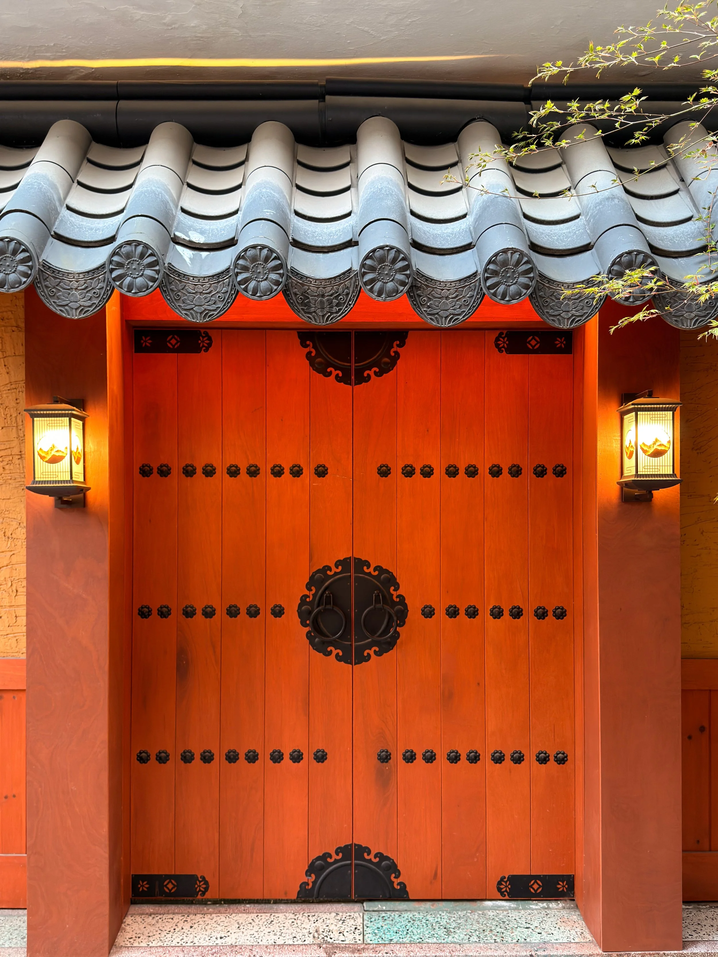 Traditional Asian-style wooden door with black decorative fittings, flanked by two lantern-style wall lights, and an ornate roof with gray tiles.