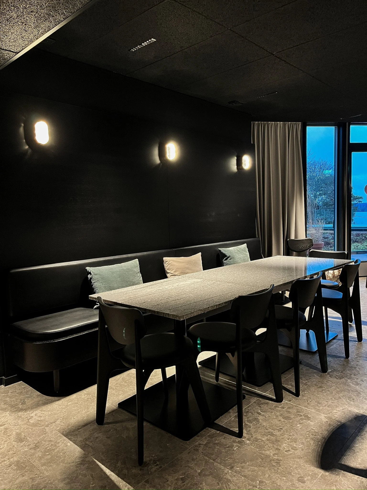 Modern dining room with a large table, six chairs, and a black cushioned bench with pillows. Wall-mounted lights on a dark wall and a window with curtains showing a view of trees outside.