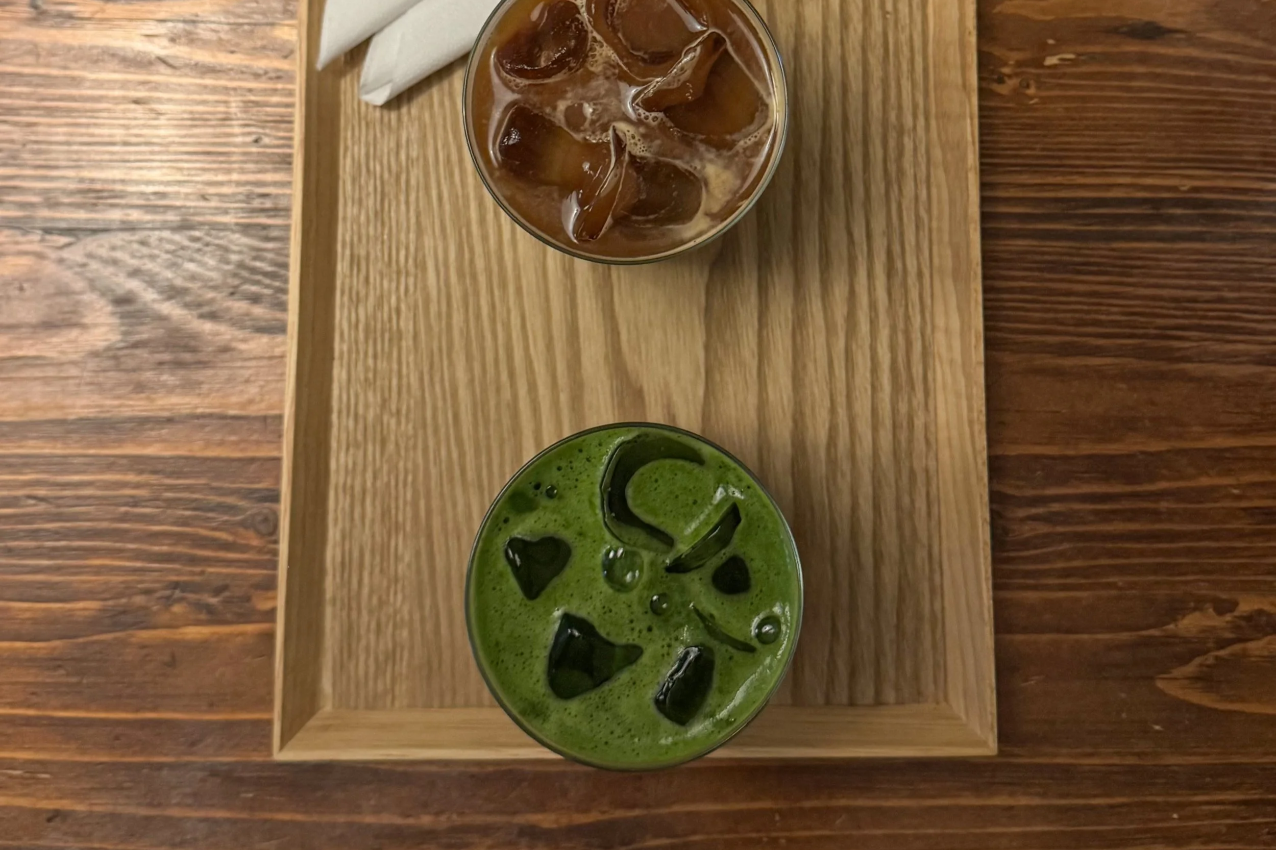 A top view of two glasses on a wooden tray, one filled with iced coffee and the other with iced matcha green tea, both with ice cubes.