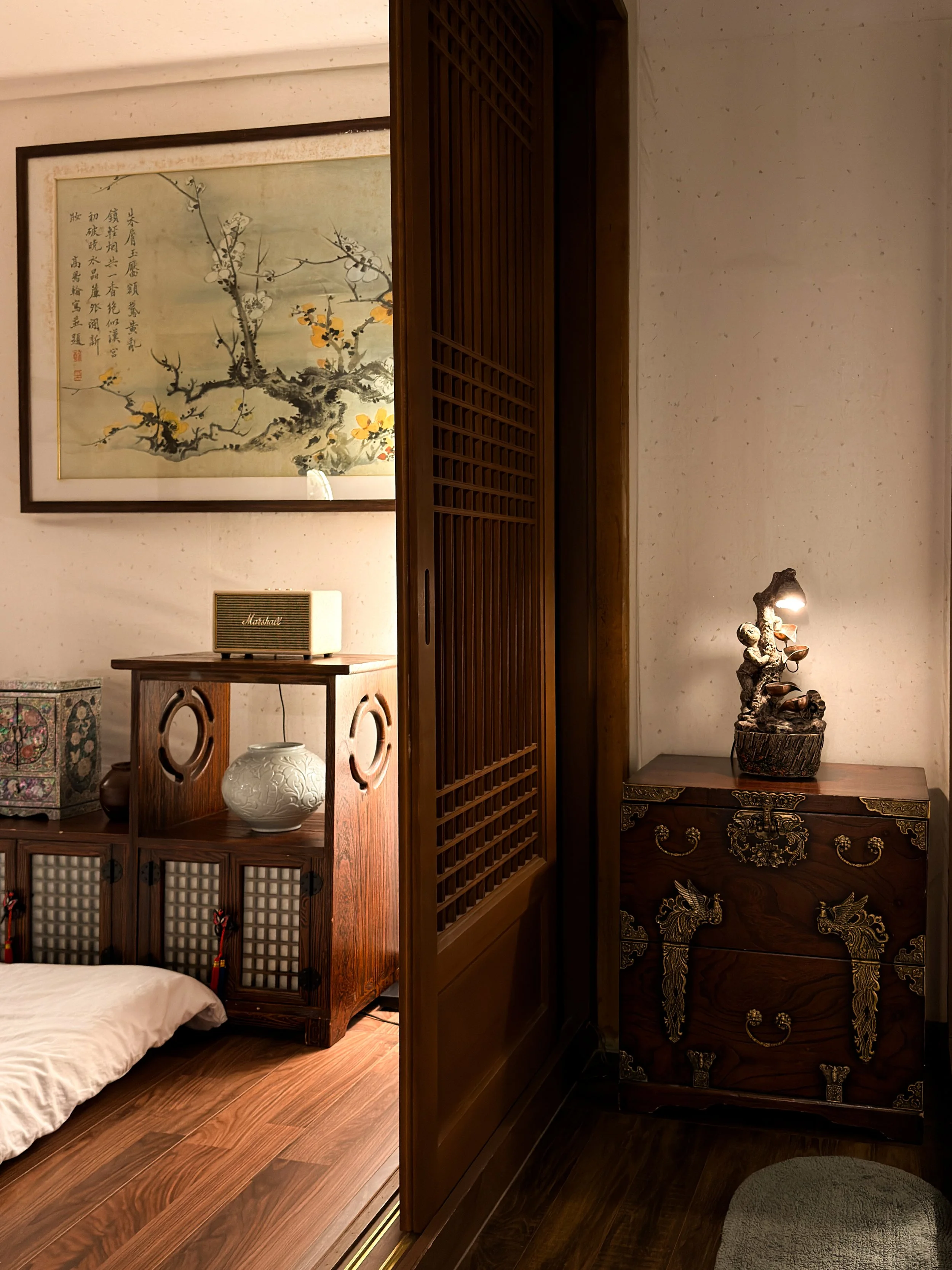 A room with Asian-style decor, showing a wooden sliding door, a bed with white bedding, Asian artwork on the wall, a small bookshelf with decorative items including a white vase, and a carved wooden chest with a small lamp on top.