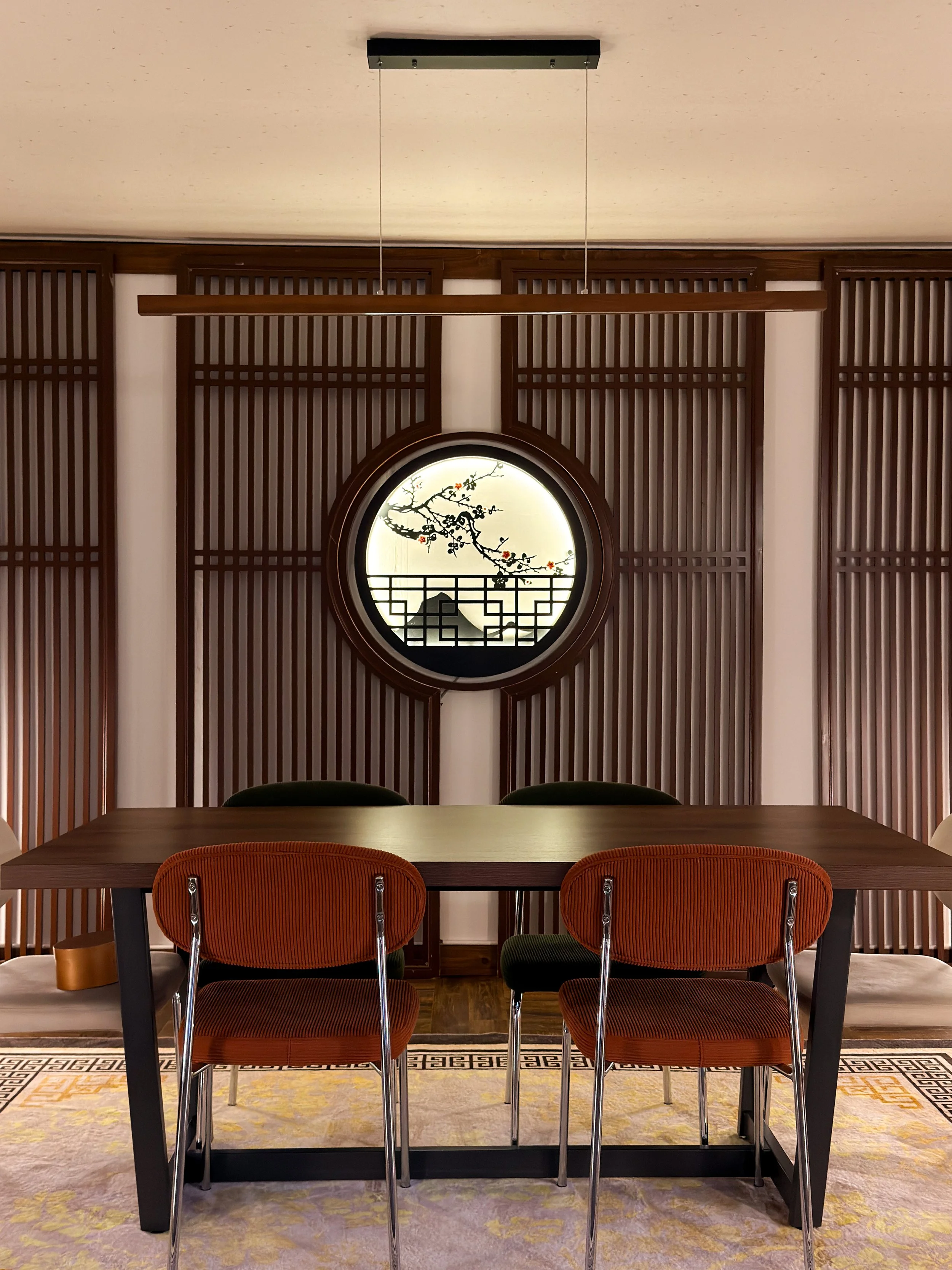 A modern dining room with wooden paneling, a round window with a floral design, and a wooden table with three striped chairs.