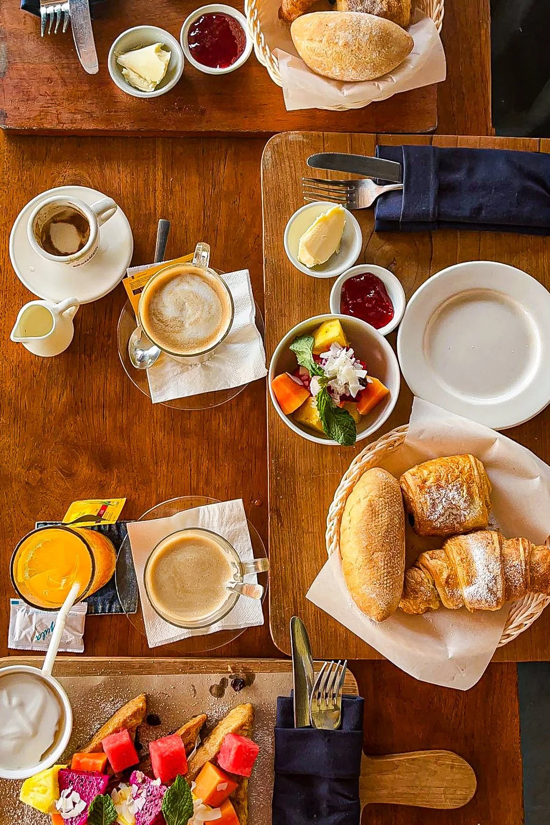 Breakfast table with bread rolls, croissants, fruit salad, coffee, tea, juice, butter, jam, and a small salad.