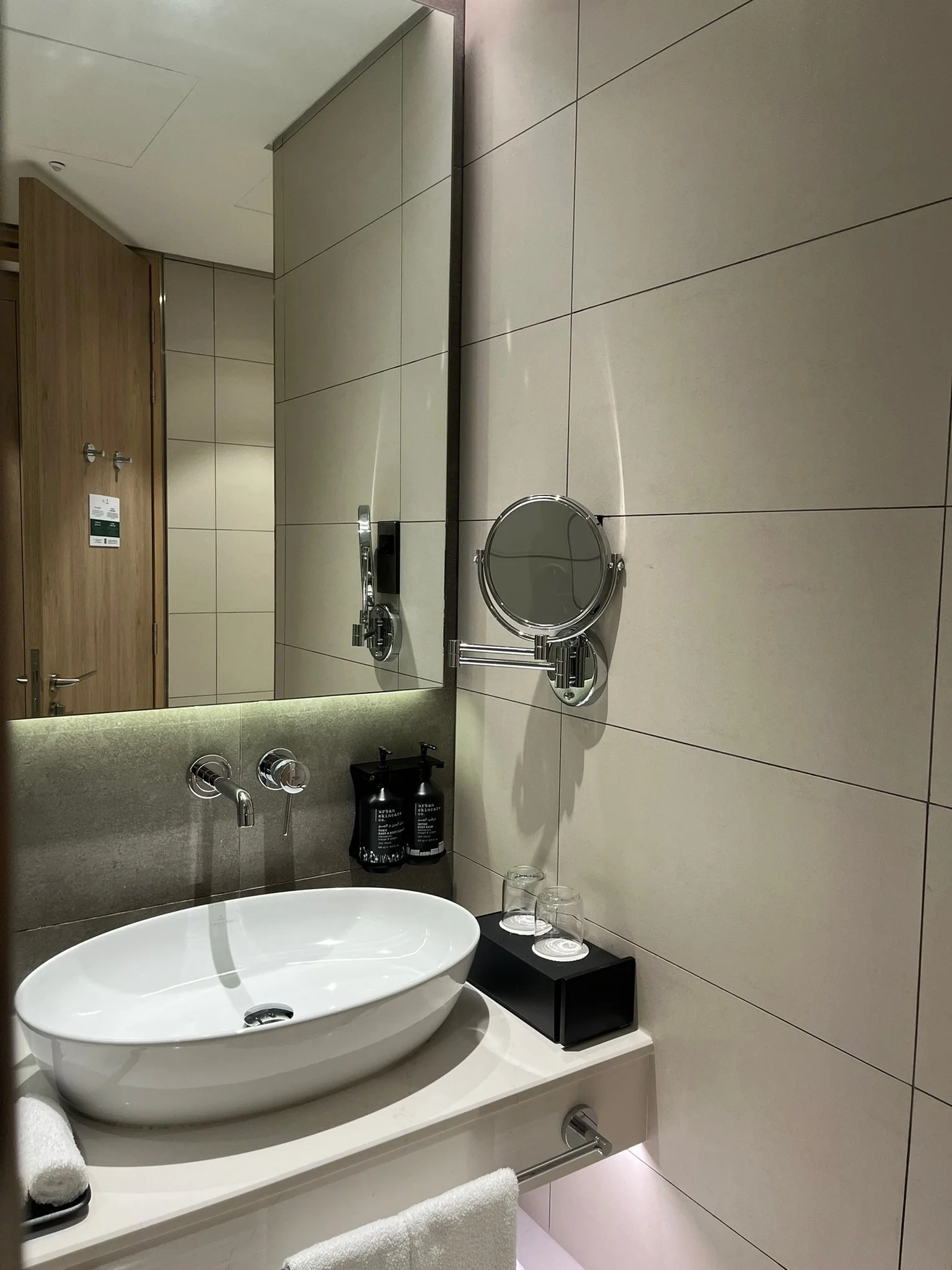 A modern hotel bathroom with a white oval sink, a large mirror, a mounted magnifying mirror, two water bottles, and two glasses on a black tray on the counter. There is a towel hanging on a bar below the sink, and beige tiled walls.
