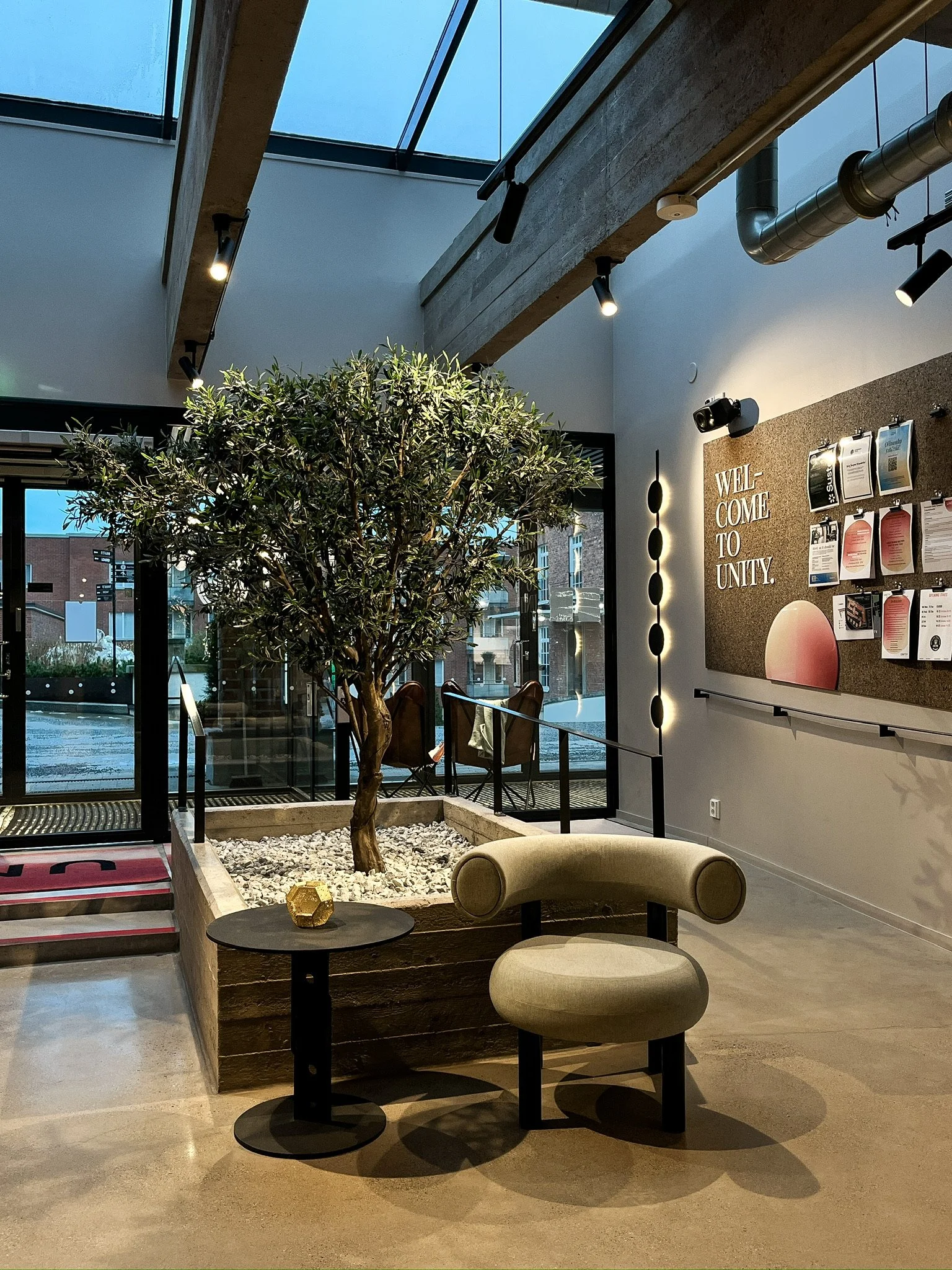 Interior of a modern lobby with a potted tree, a beige curved chair, a black side table with a gold decorative object, a bulletin board with papers, a welcome sign, and large windows letting in natural light.