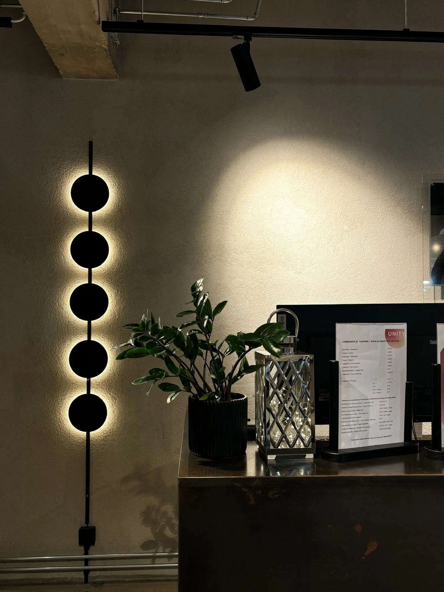 A modern interior scene with a decorative wall light consisting of four black circles vertically aligned, casting soft light on a light-colored wall, next to a wooden counter with a black potted plant, a metal lantern, and a white menu stand.
