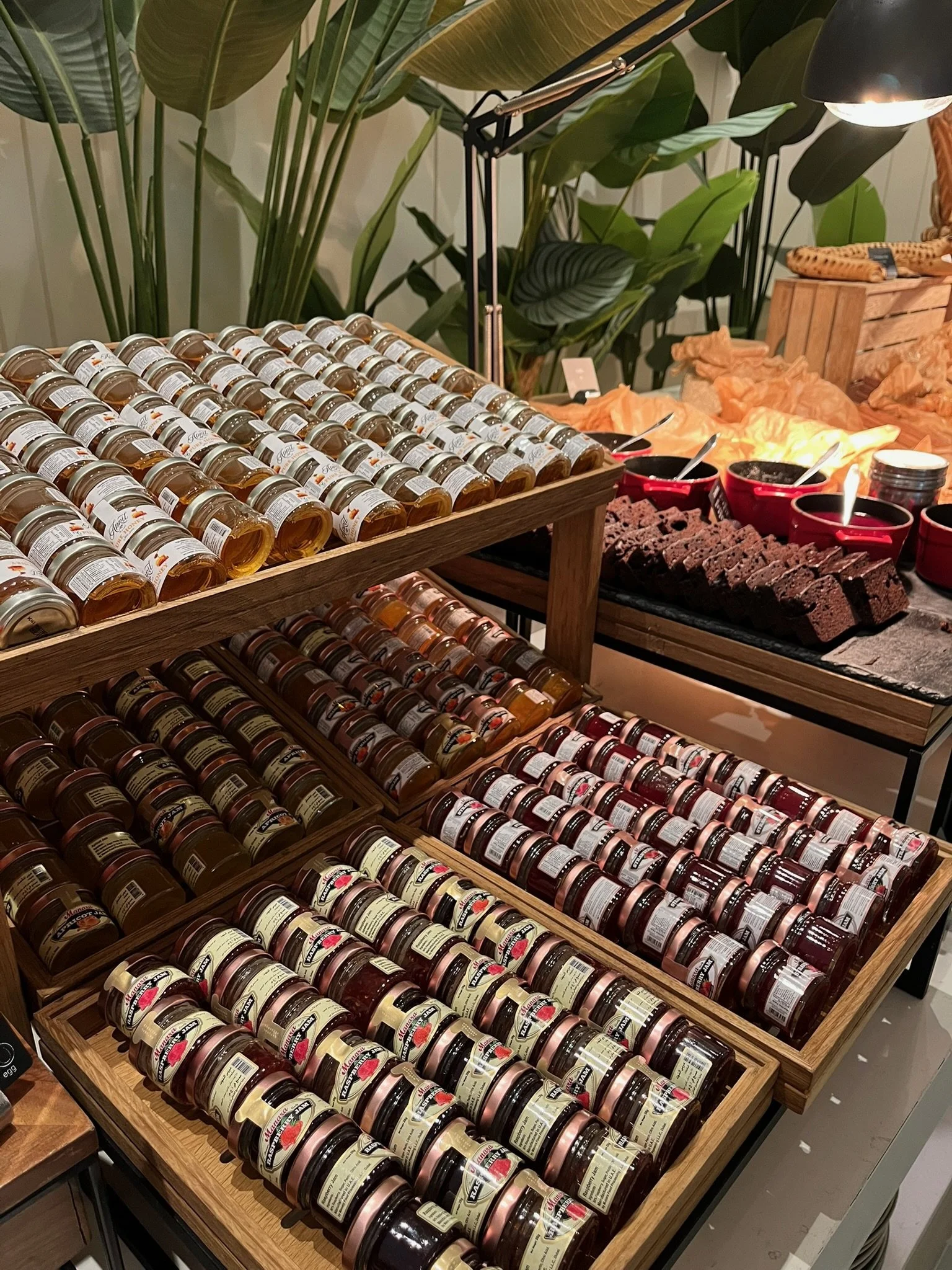 Display of various jars of jam on wooden shelves in a store, with jars organized by flavor or type. In the background, there are bowls of what appears to be chocolate spread and other food items on a table, with green leafy plants and a desk lamp.