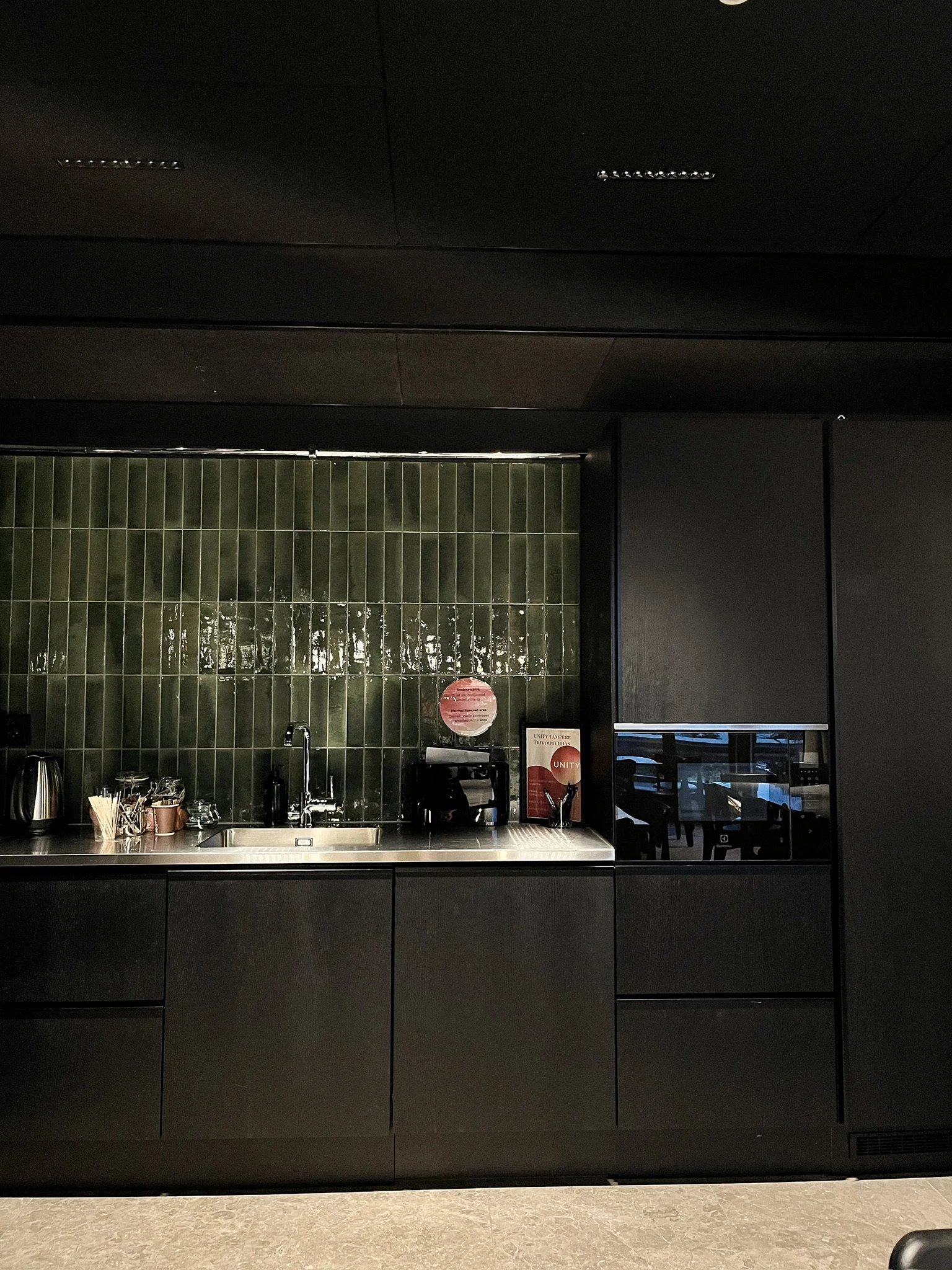 A minimalist black kitchen with dark cabinets, a stainless steel sink, green tile backsplash, and a few small appliances and items on the counter.