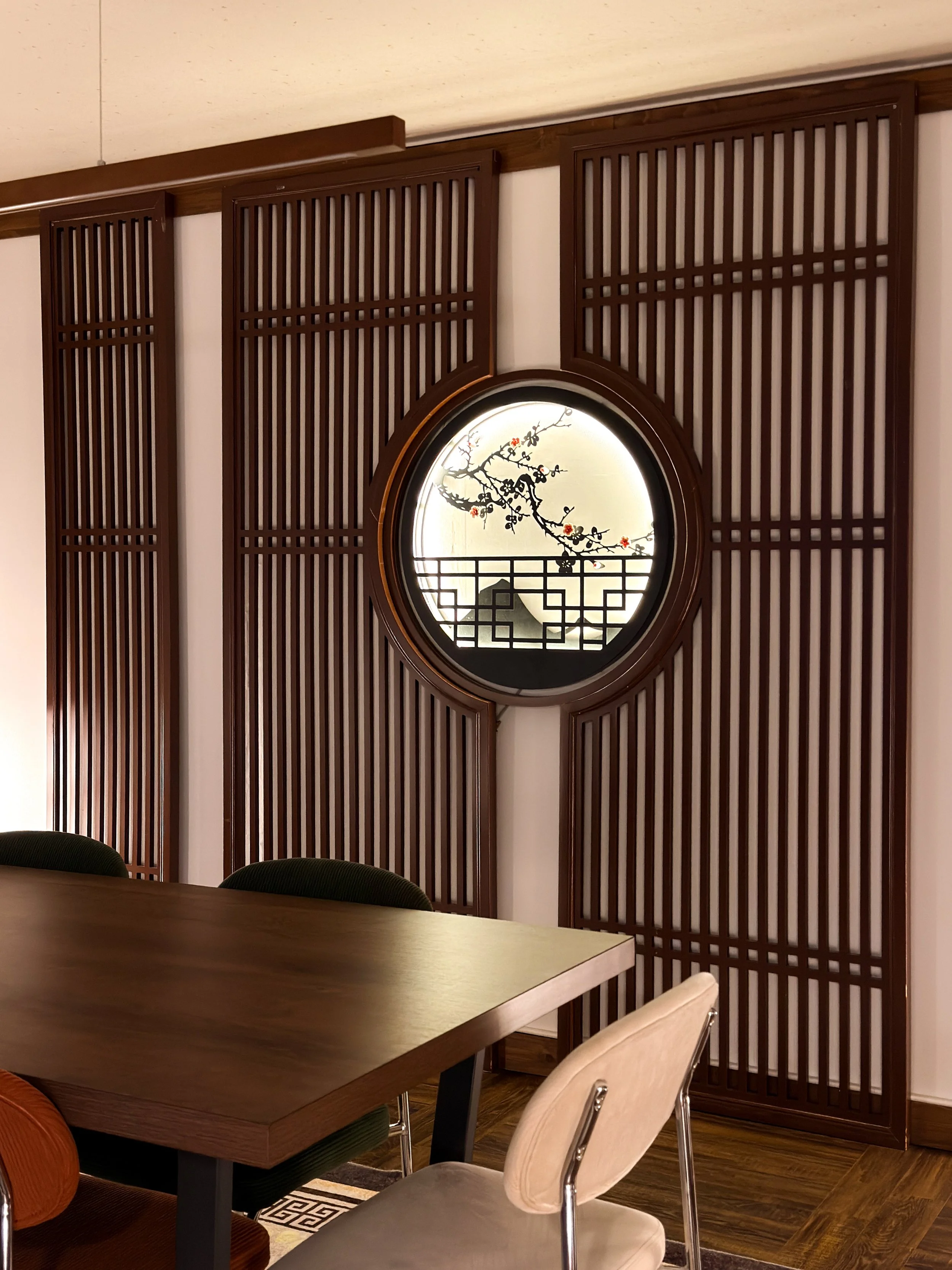 Interior of a room with a wooden table and chairs, featuring a decorative round window with an Asian-style design and a branch with blossoms.