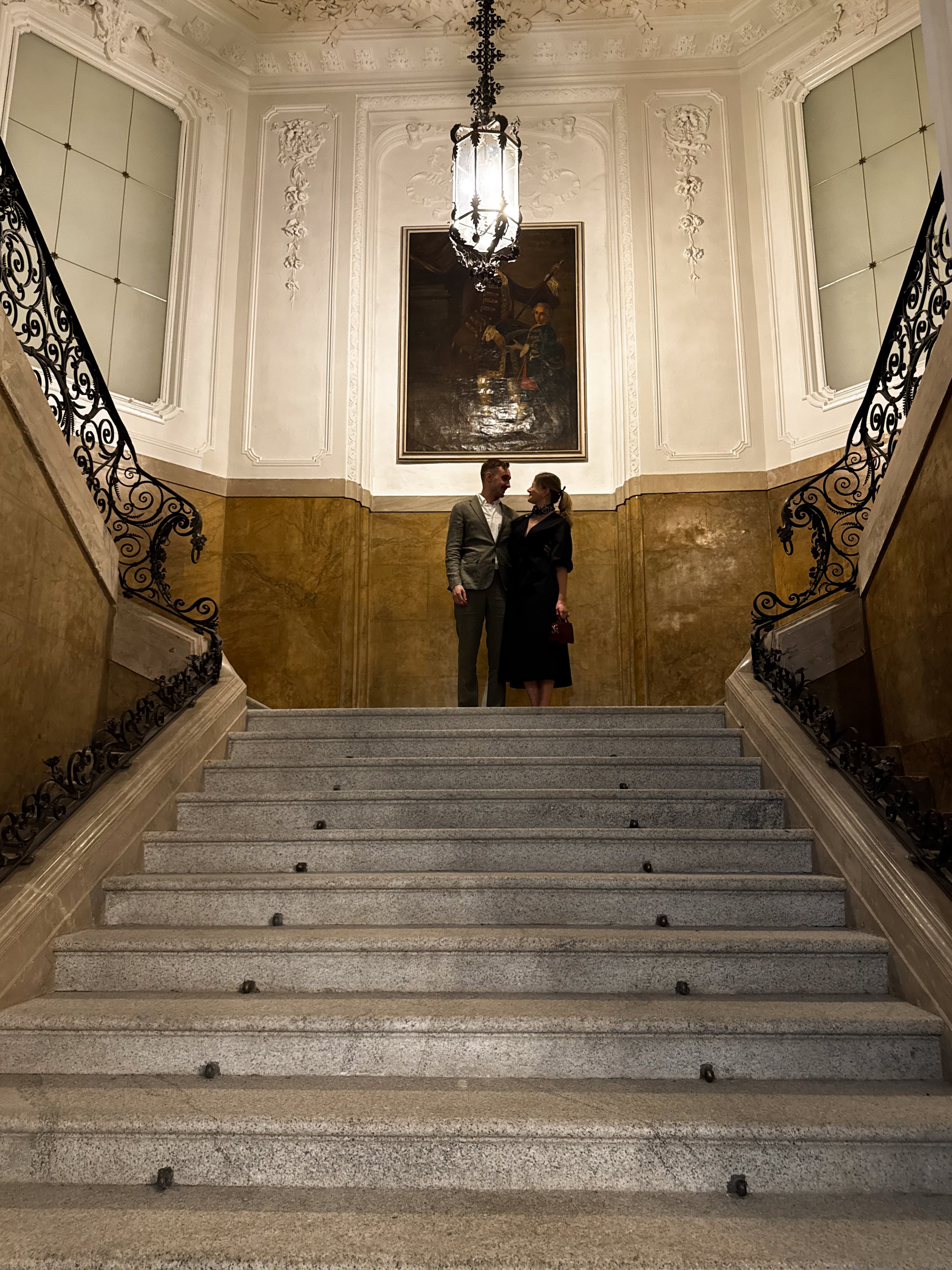 A man and a woman standing and facing each other at the top of a grand staircase in an ornate hallway, with a large painting and elaborate molding on the walls behind them.