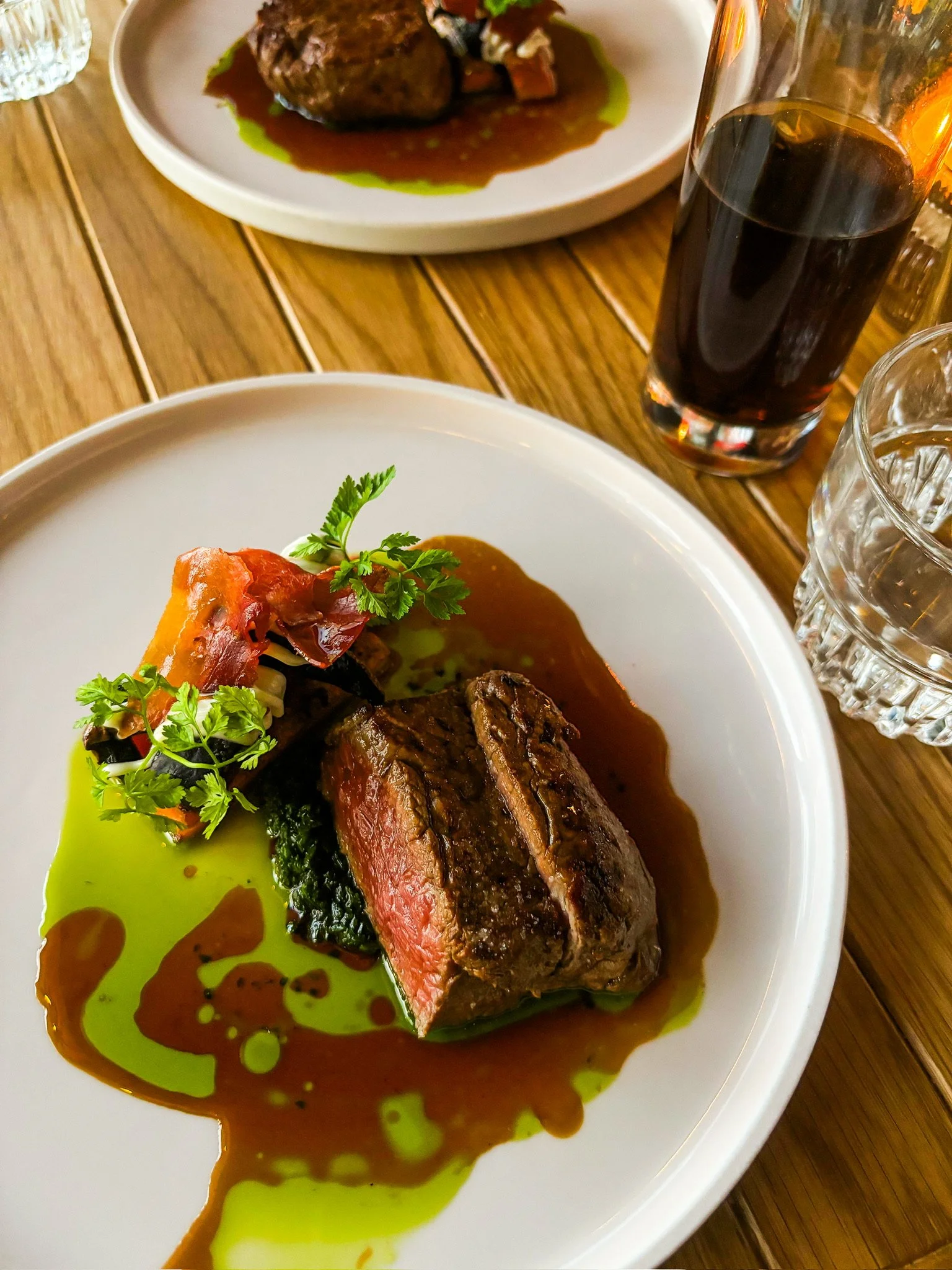 A plate with a piece of medium-rare filet mignon steak, garnished with herbs, with green and brown sauces on a white plate. In the background, a glass of dark soda, a glass of water, and another plate with a meat dish are visible on a wooden table.