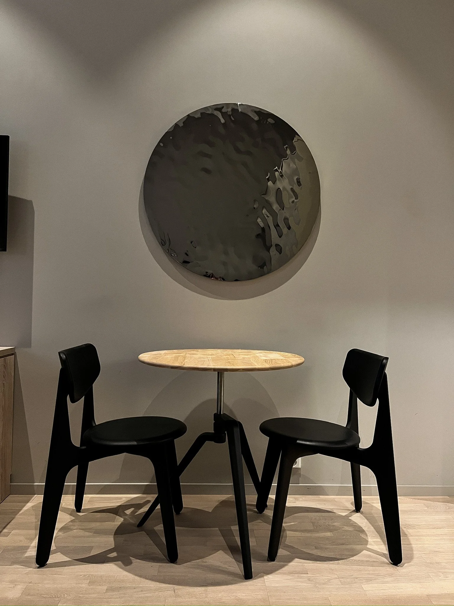 A small round wooden table with black chairs, placed in front of a white wall with a large circular mirror reflecting an abstract image.