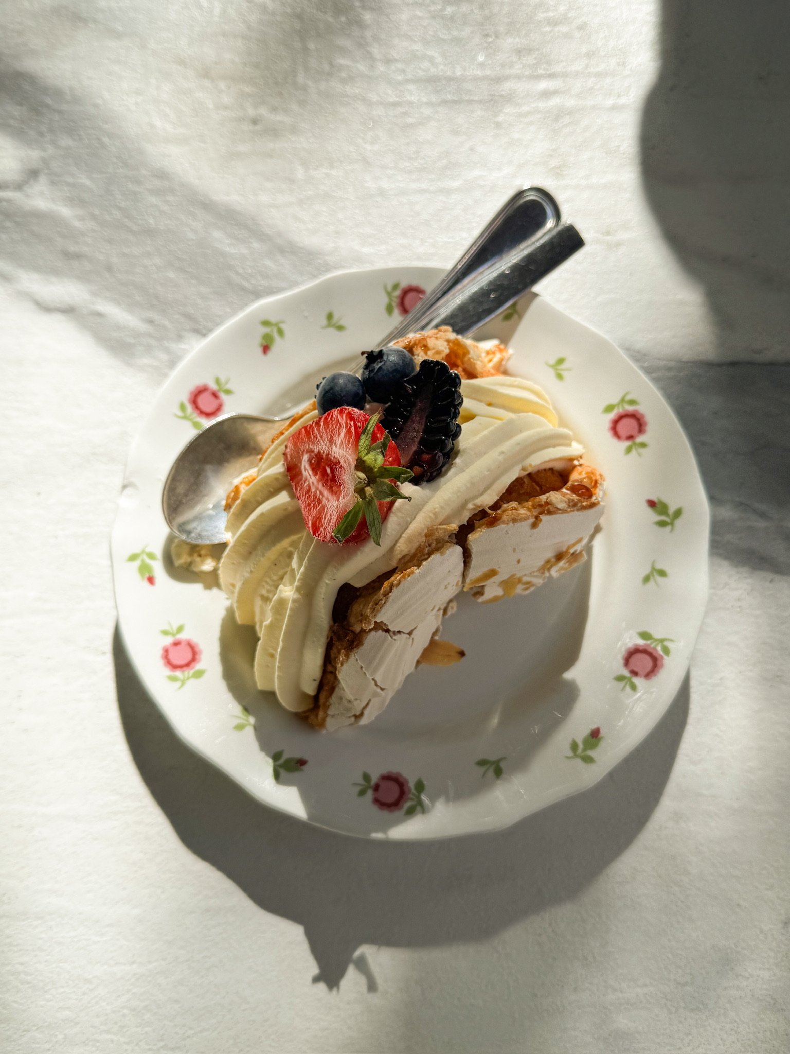 Slice of layered cake with white frosting, topped with fresh strawberries, blackberries, and blueberries, served on a floral-patterned plate with a spoon and fork.