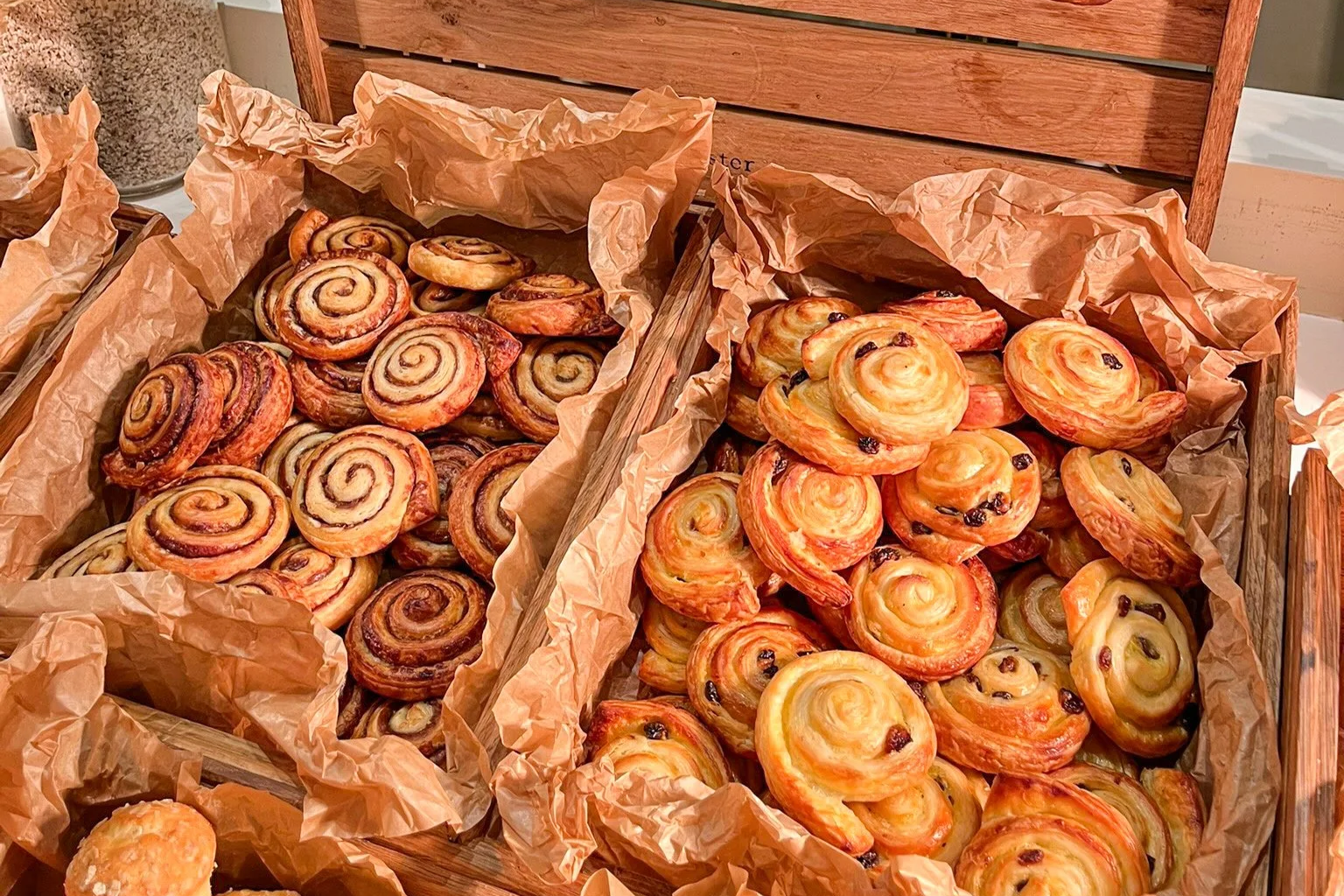 Assorted baked pastries including cinnamon rolls and Danish pastries with raisins, displayed in baskets lined with brown parchment paper.