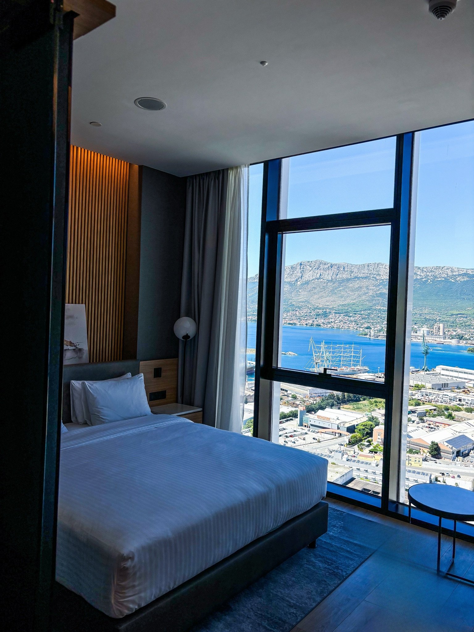 Hotel room with a large window view of a city, water, mountains, and harbor with ships during daytime.