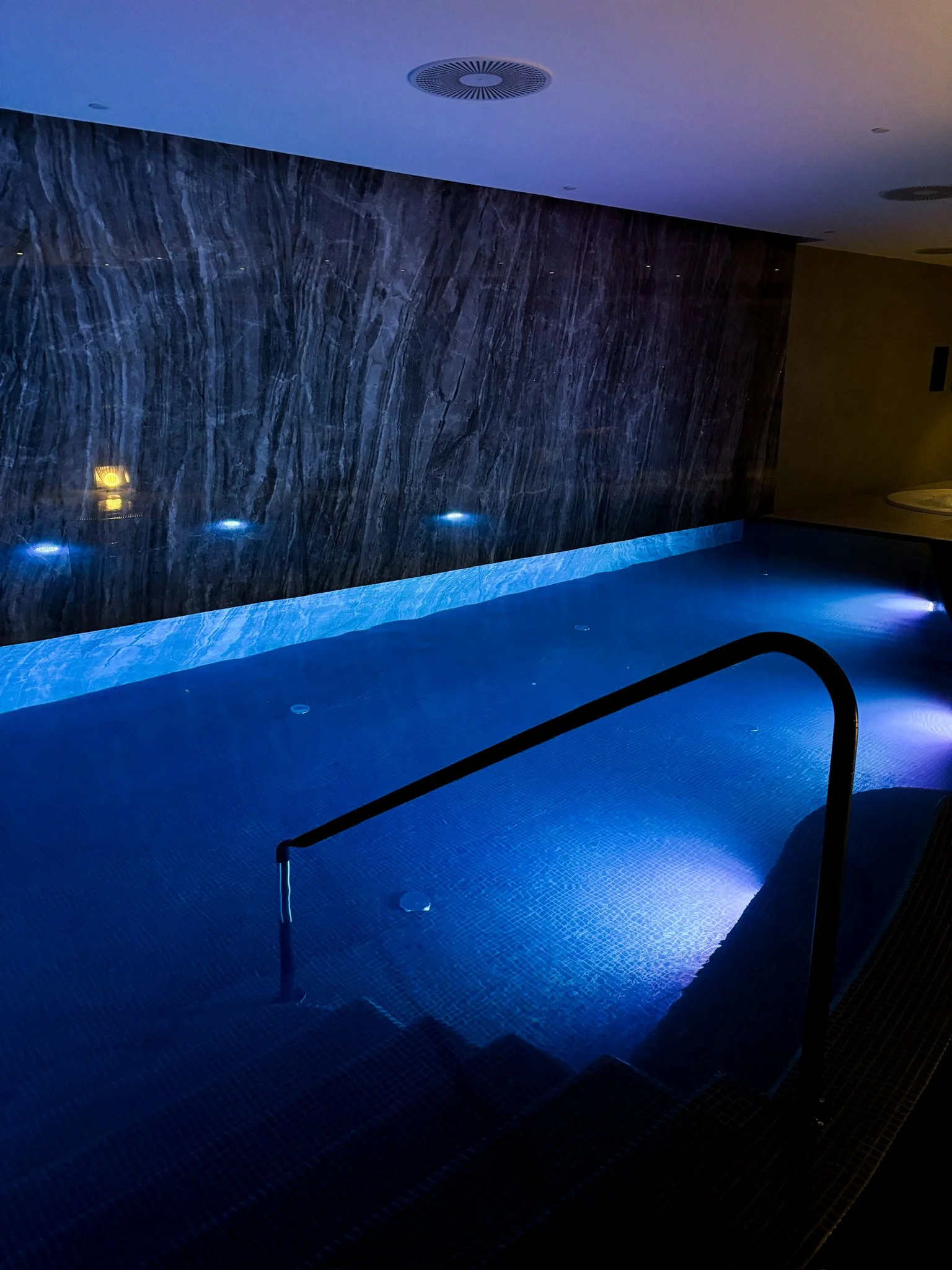 Indoor swimming pool with blue lighting, dark marble wall, and steps with a black handrail.