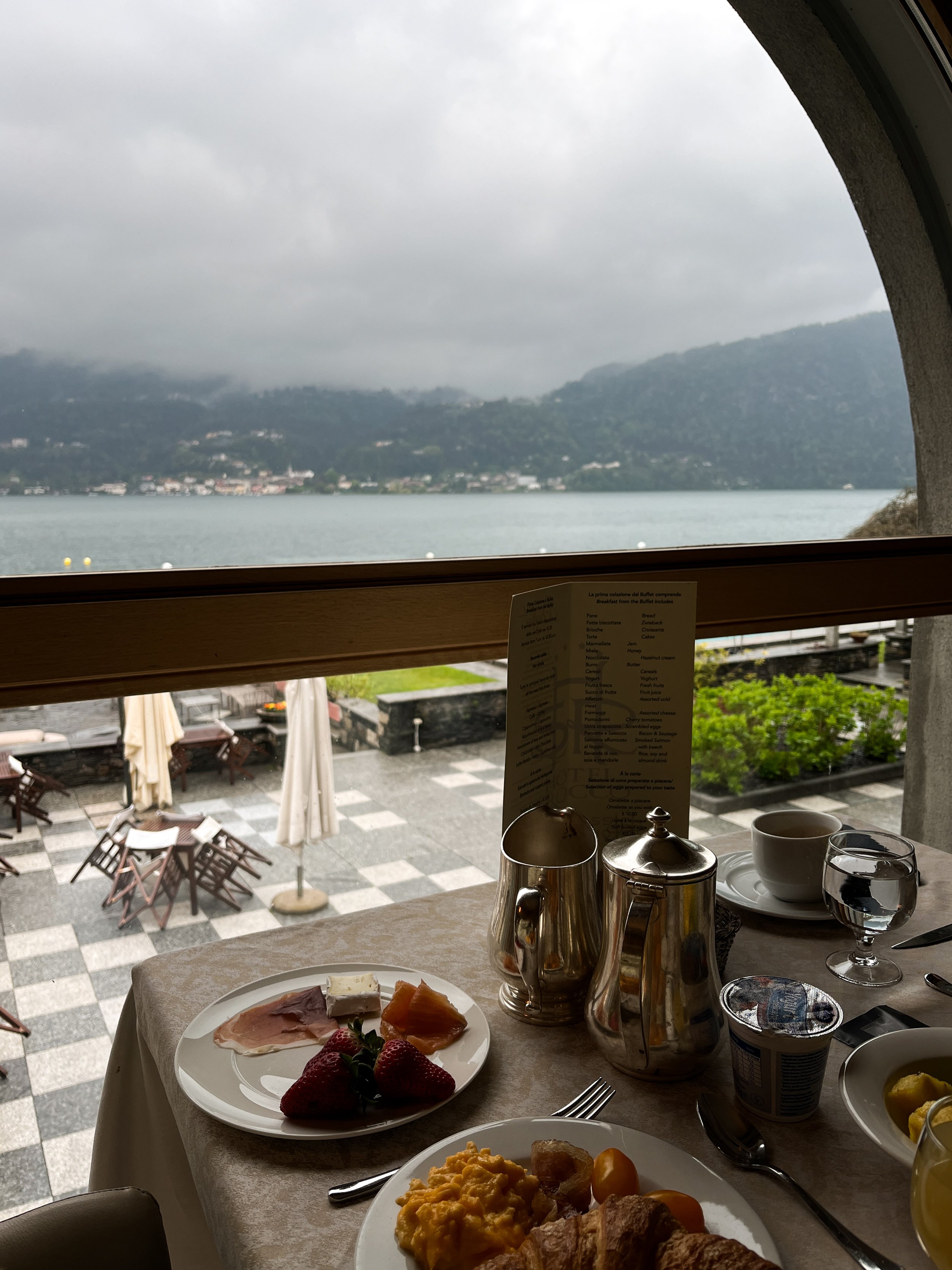 A breakfast table with scrambled eggs, croissants, strawberries, cheese, sliced ham, and smoked salmon, set near a window overlooking a lake and mountains on a cloudy day.