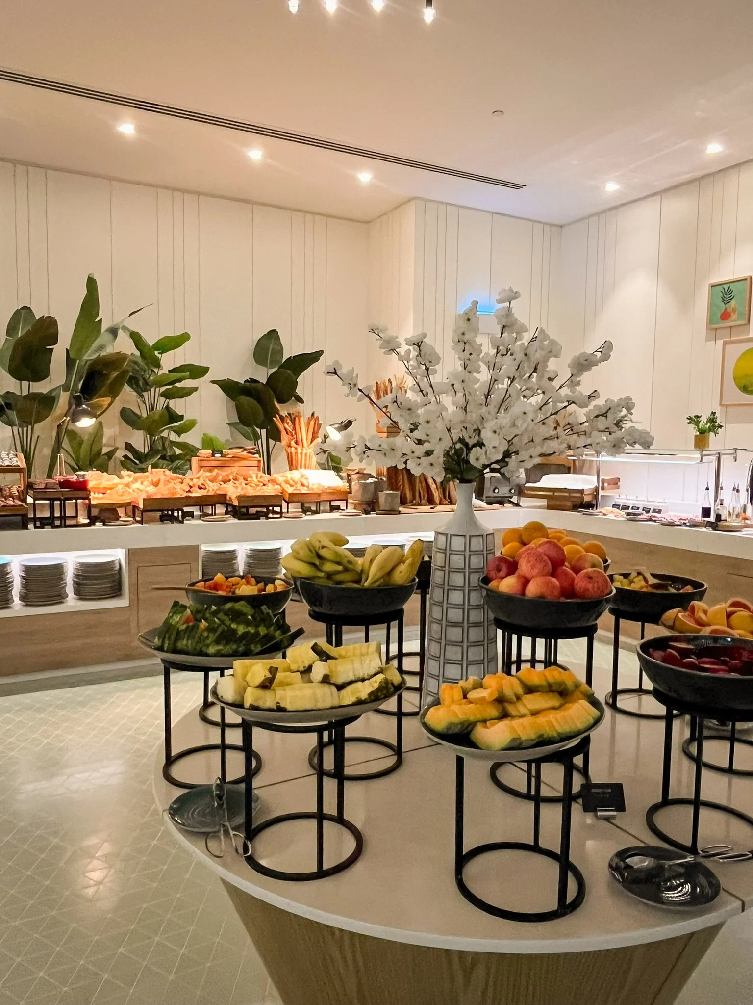 Fruit display with bananas, peaches, honeydew, and watermelon in black bowls on round stands, with a white floral centerpiece in a tall vase and a buffet table in the background in a buffet or dining area.