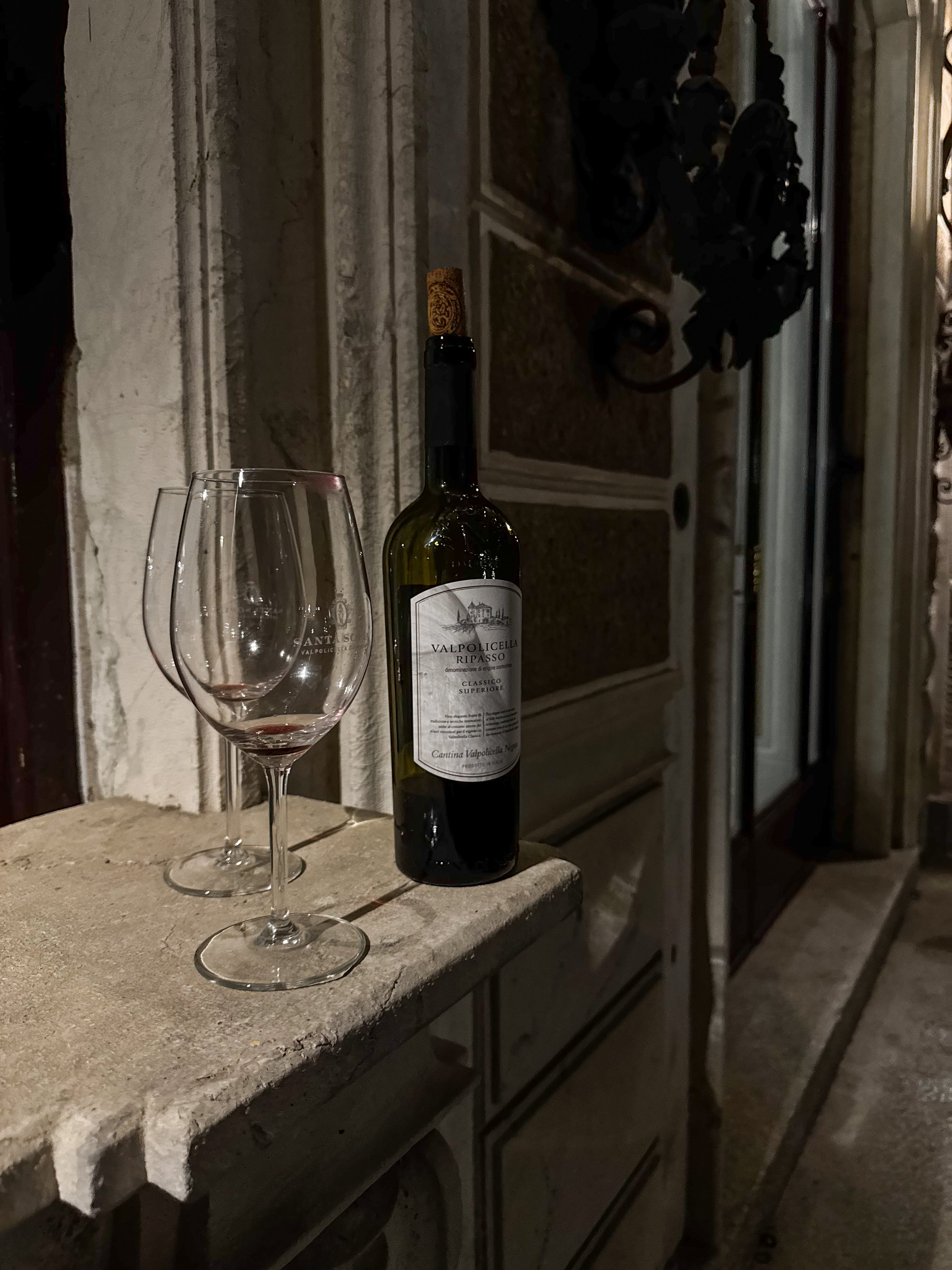 A partially filled wine glass, a full wine bottle, and a second empty wine glass on a stone ledge outside a building, with a door and wall in the background.
