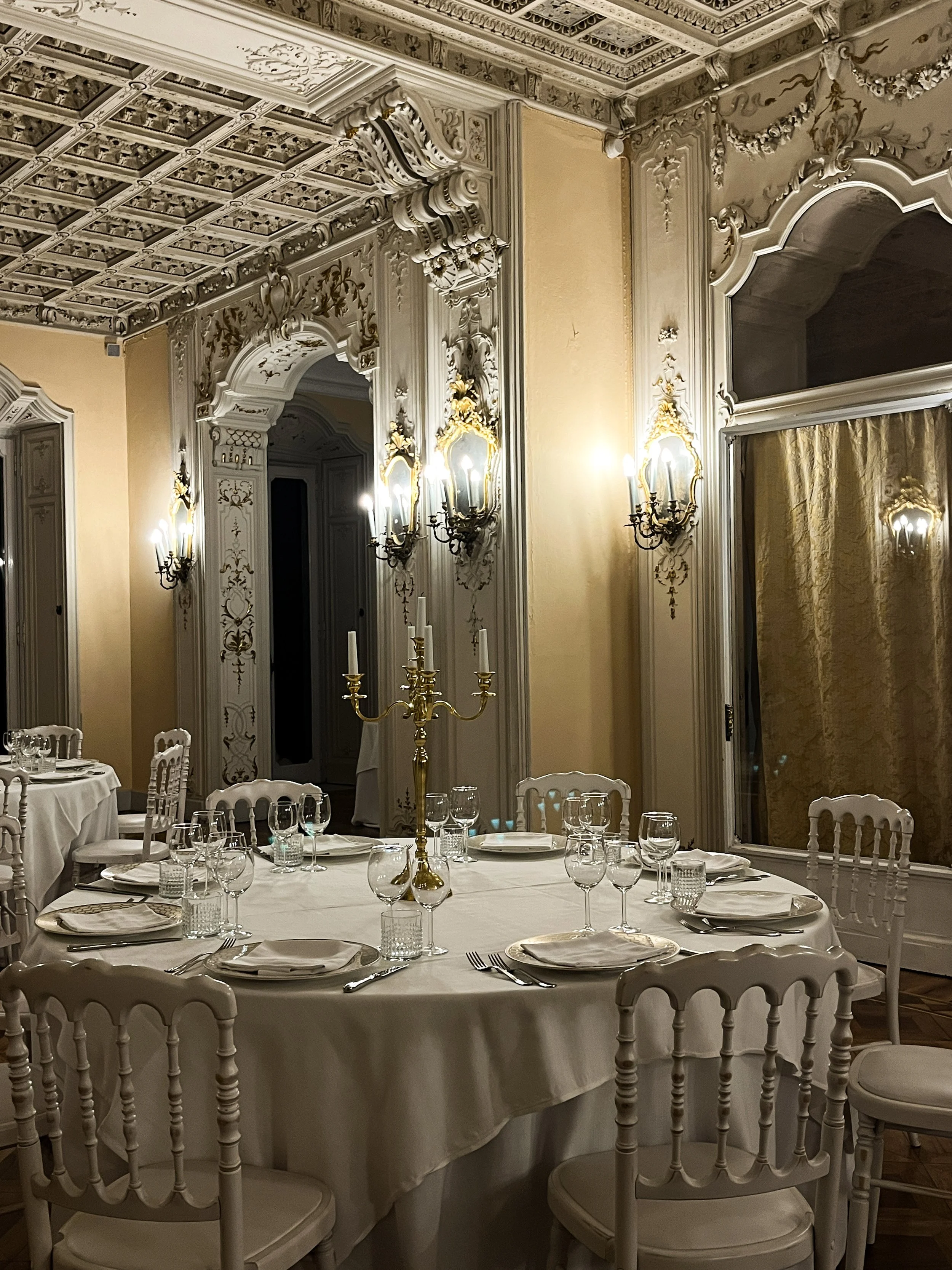 Elegant dining room with a large round table set with white tablecloth, plates, silverware, wine glasses, and a gold candelabra centerpiece. The room features ornate white molding, chandeliers, and gold curtains.