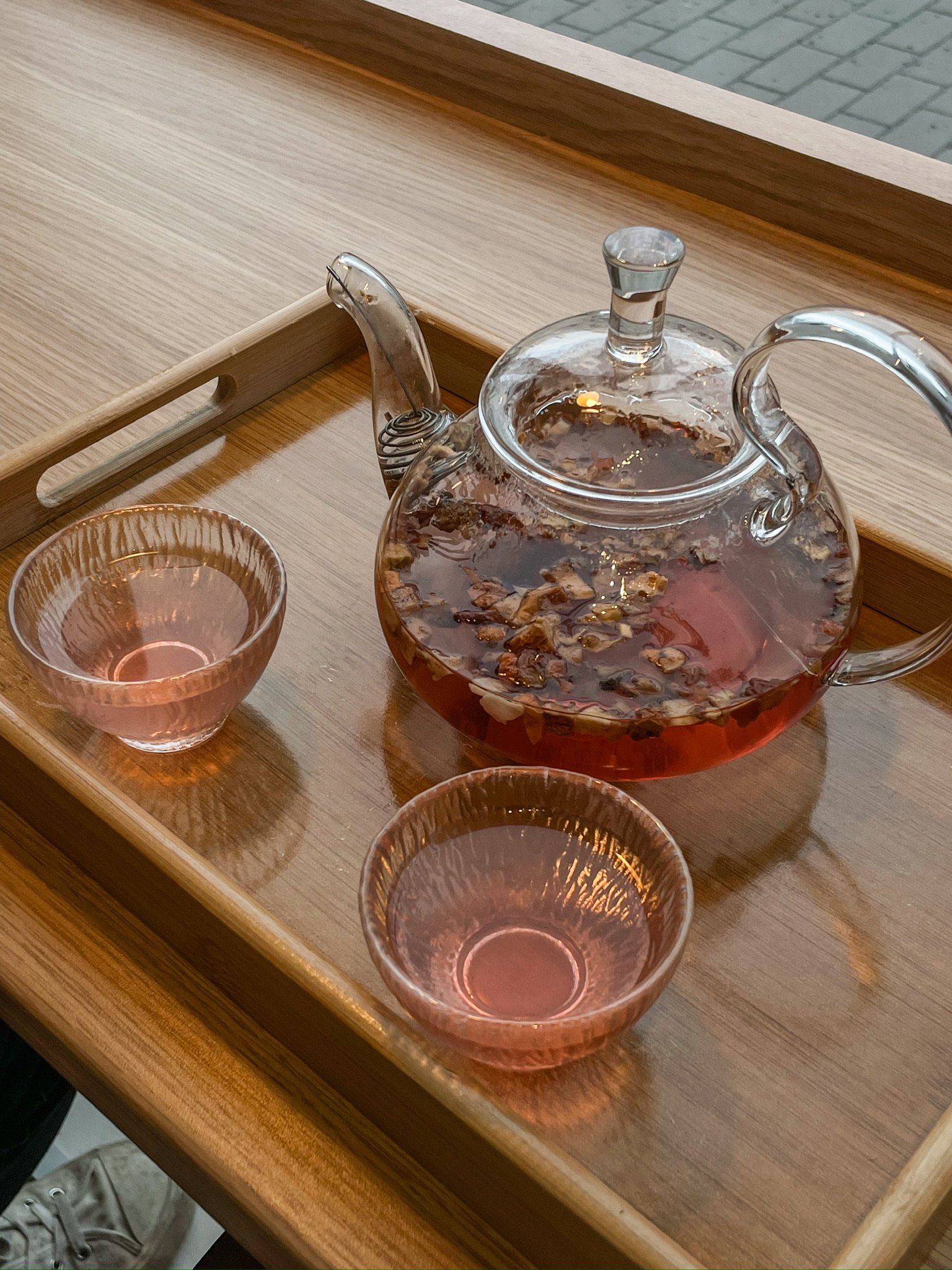 A glass teapot with a metal spout and handle, filled with tea and floating tea leaves, sits on a wooden tray. Two pink glass cups filled with tea are also on the tray.