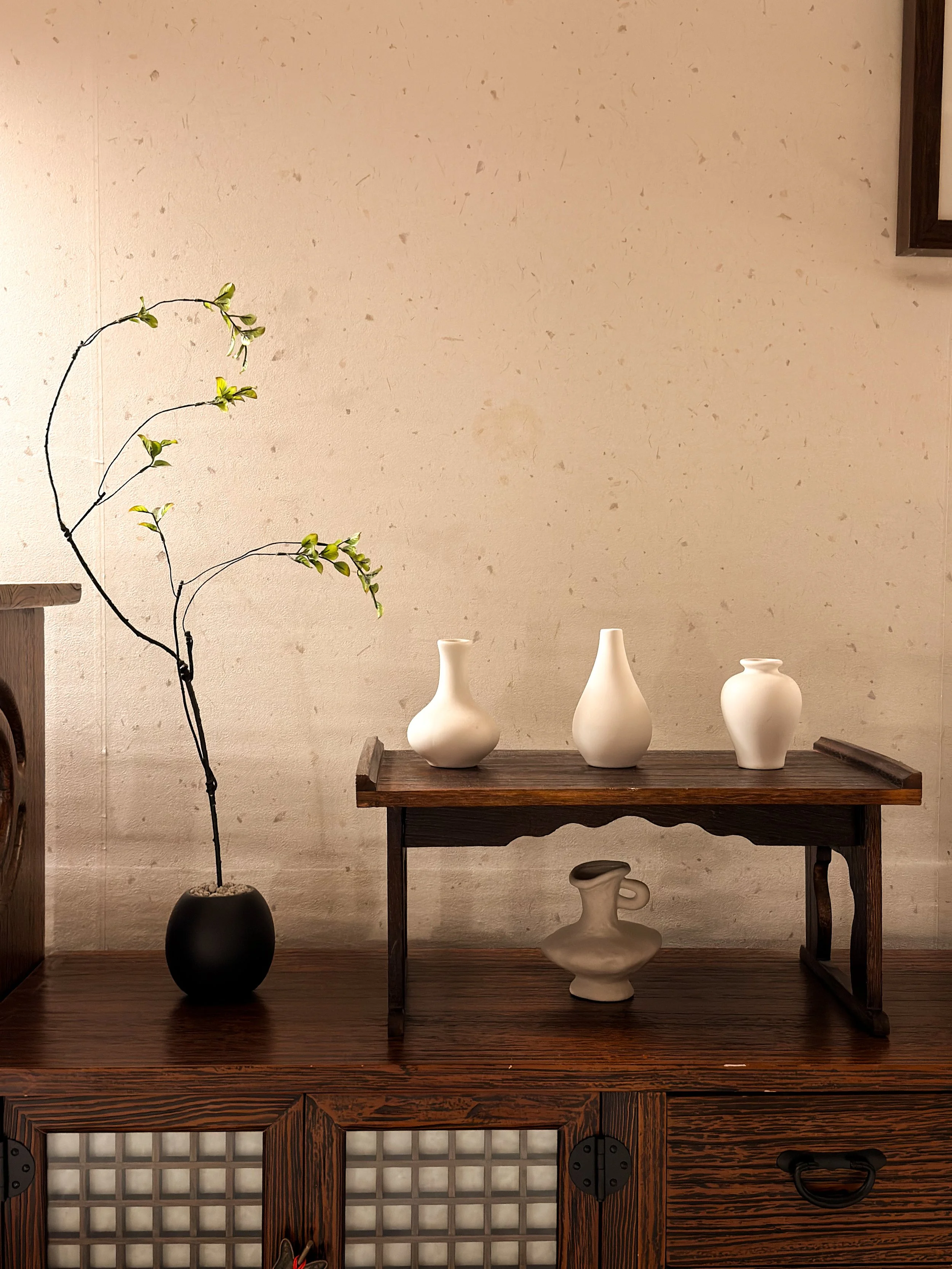 Decorative display of three white ceramic vases on a dark wooden table, with a single black vase holding a thin branch with green leaves, and a smaller ceramic vase placed underneath on a wooden cabinet.
