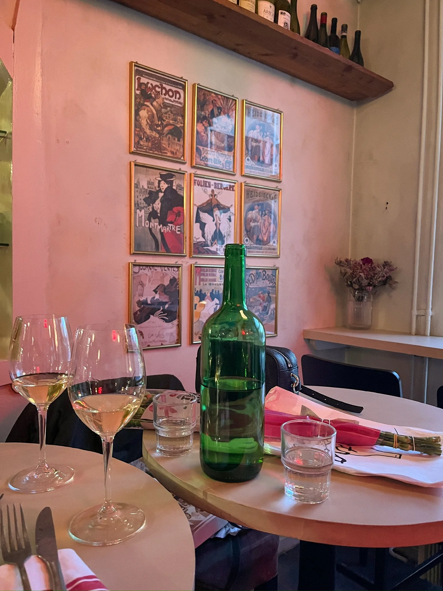 A dining table set with two wine glasses, a green bottle, a glass of water, a plate of food, a pink napkin wrapped around a fork and knife, and another glass of water. In the background, there are framed posters on the wall, a wooden shelf with wine bottles, and a vase with flowers on a windowsill.