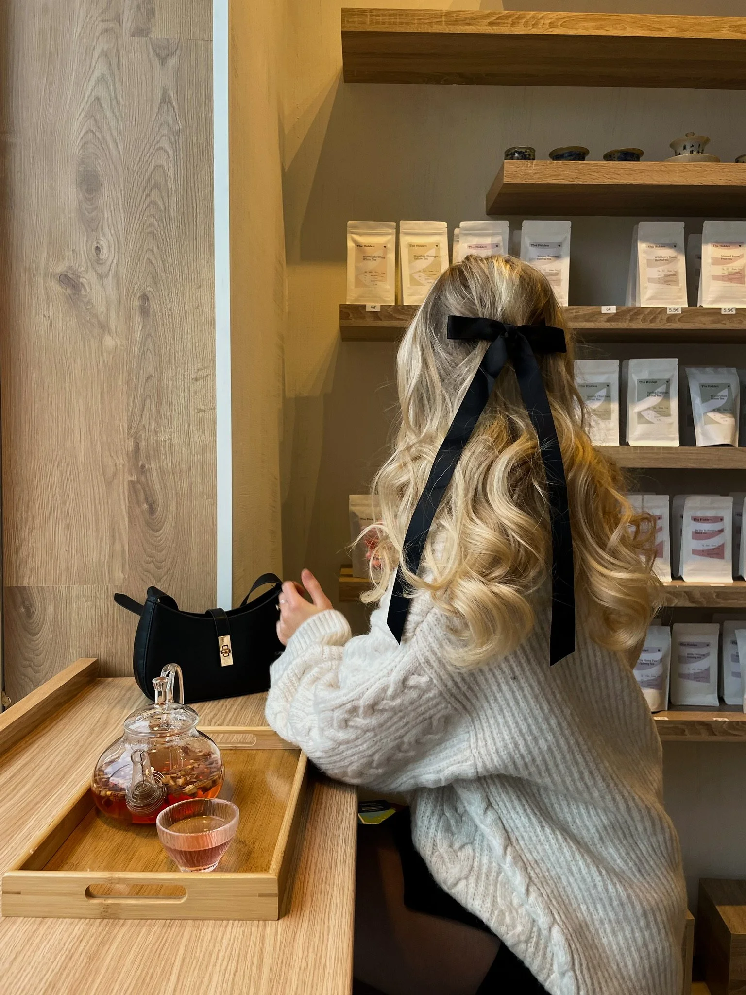 A girl with long, curly blonde hair with a black ribbon bow, wearing a beige sweater, sitting at a wooden counter in a tea shop, facing shelves of tea bags and a glass teapot with pink tea on a tray.