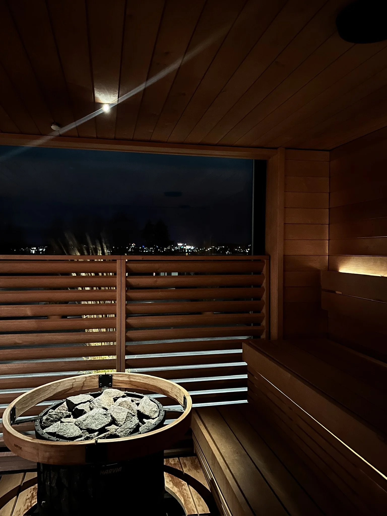 Night view of a wooden sauna interior with a bench, a circular heater with rocks, and a window showing a dark city skyline at night.