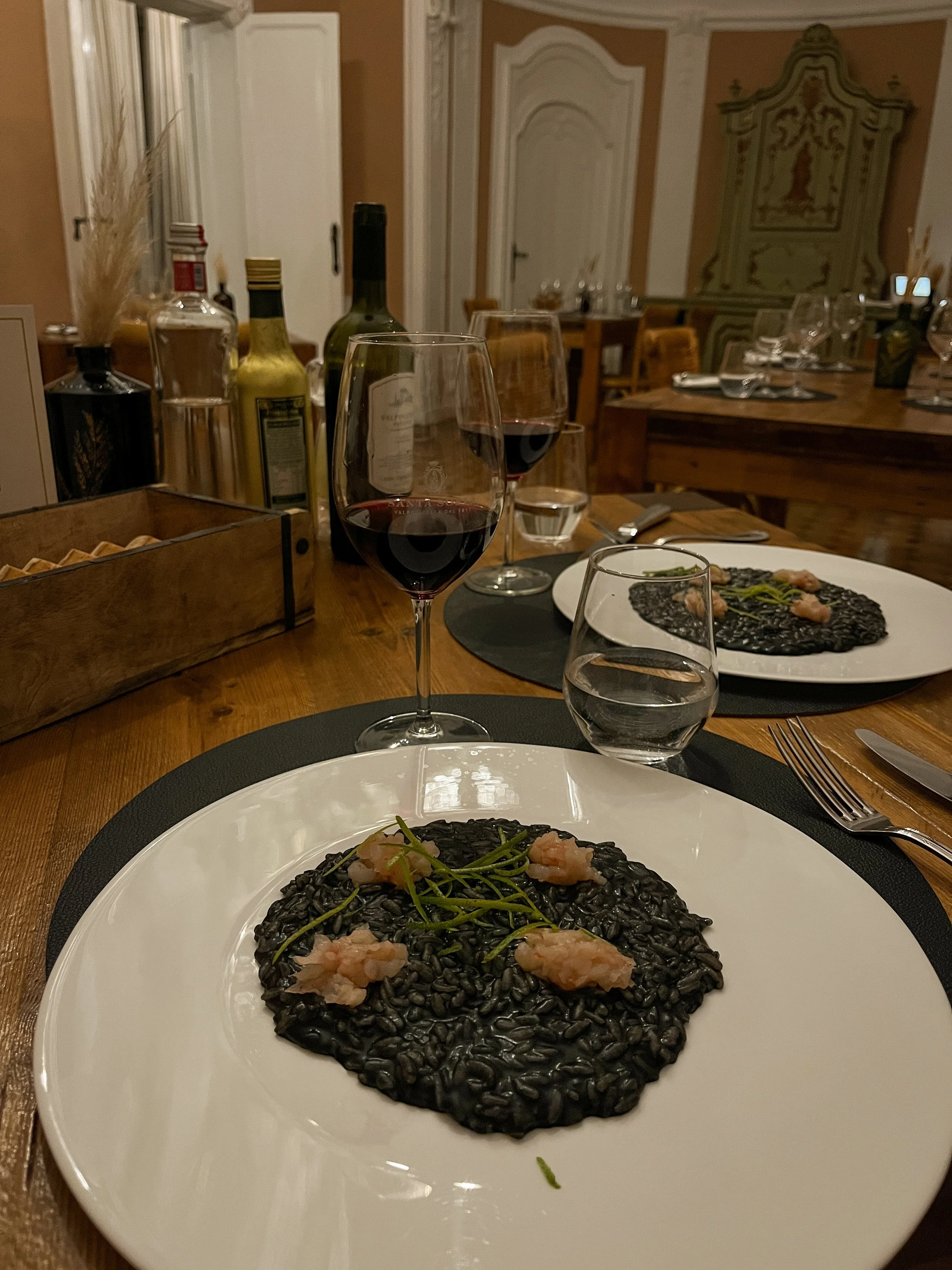 A dinner table with a white plate of black risotto topped with shrimp and green herbs, two glasses of red wine, a glass of water, and additional plates and condiments in a cozy dining room.